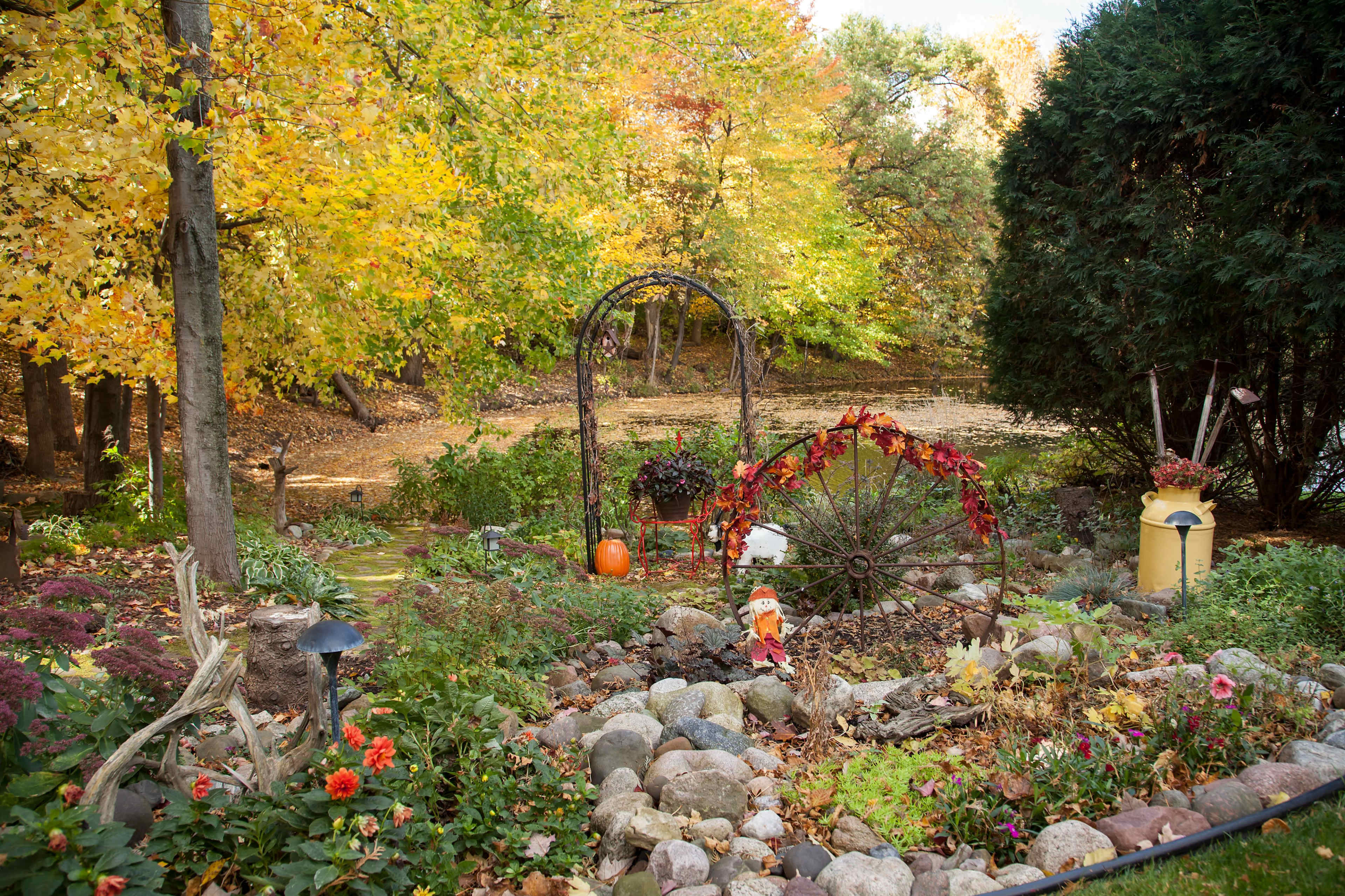 A garden features a decorative arch with fall foliage, surrounded by colorful flowers and rocks, leading to a pond in the background.
