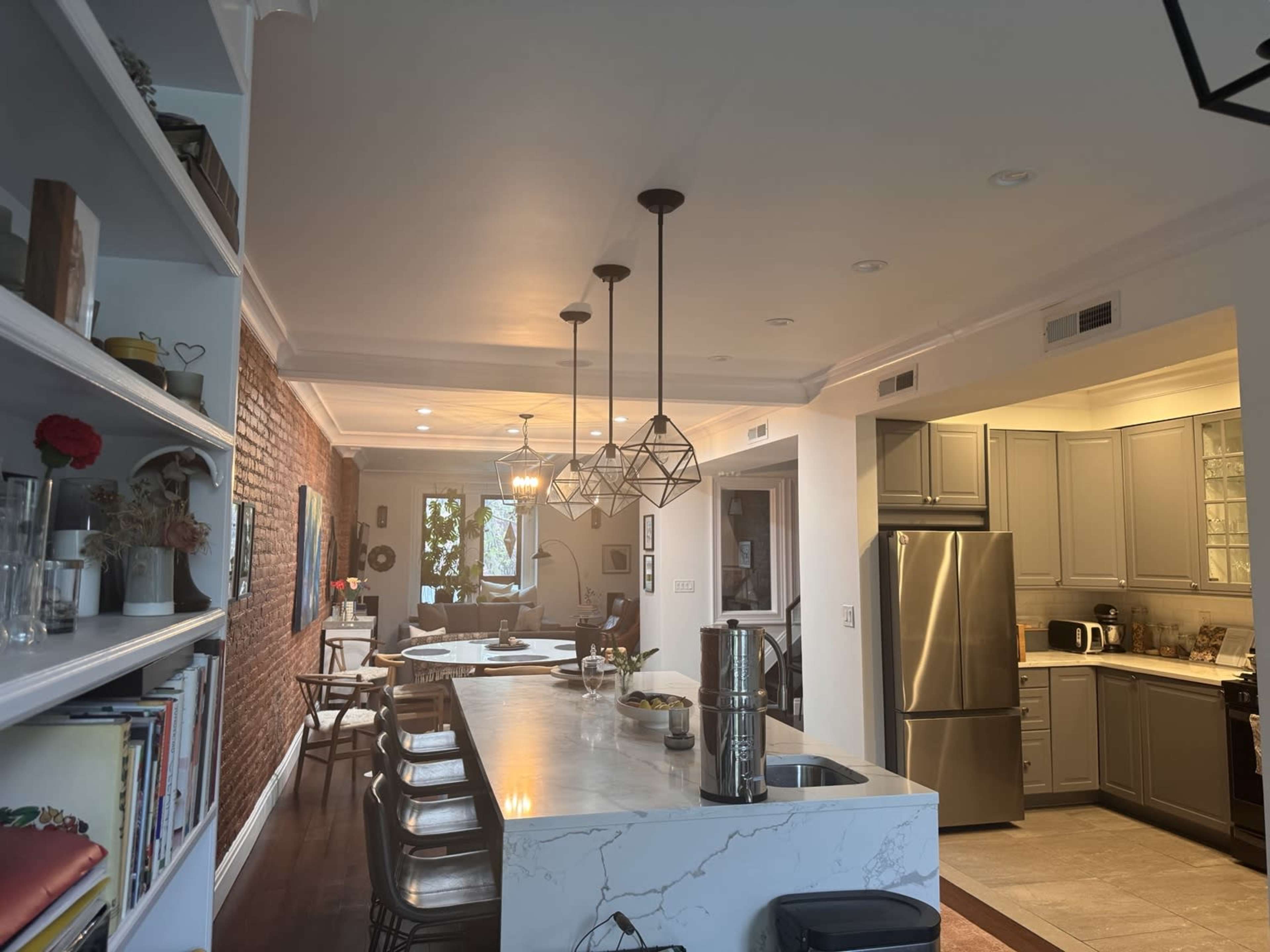 The image shows a modern kitchen and dining area with a long marble island, pendant lights, and a brick accent wall.