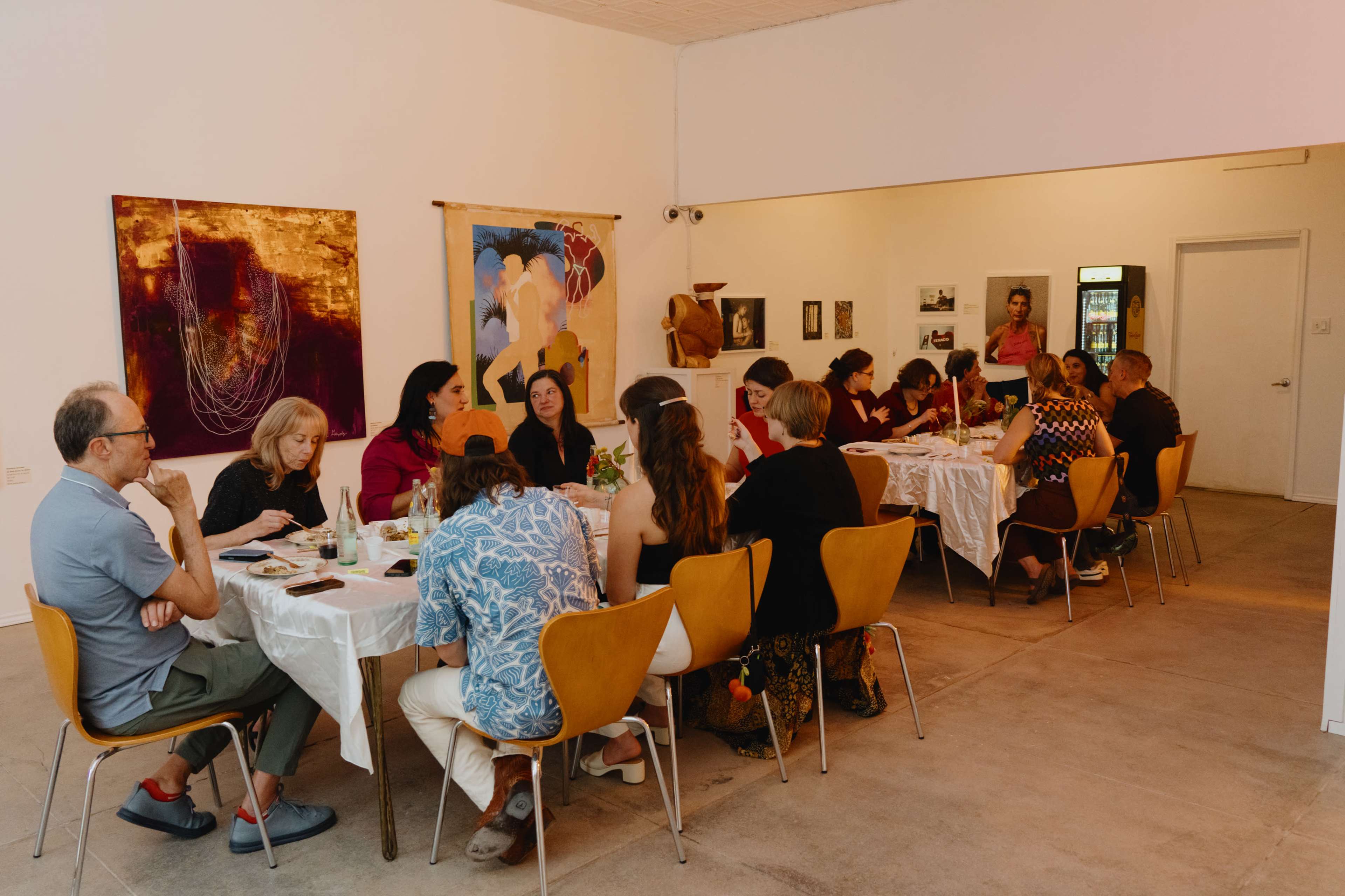 A group of people is seated at two tables in an art gallery, engaged in conversation during a dining event.