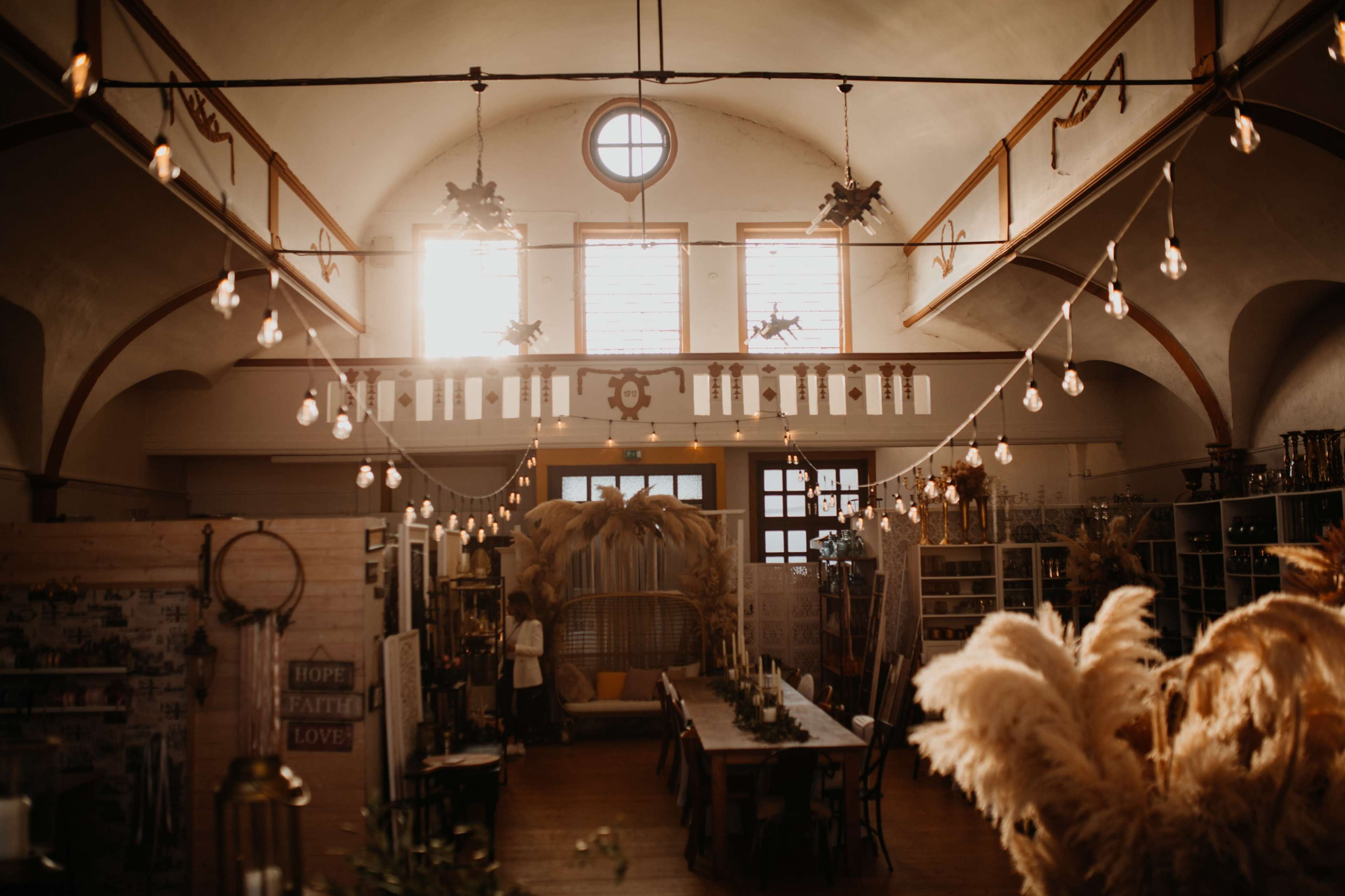 The image shows an interior space with high ceilings, soft lighting from hanging bulbs, and a long table surrounded by decorative elements and greenery.