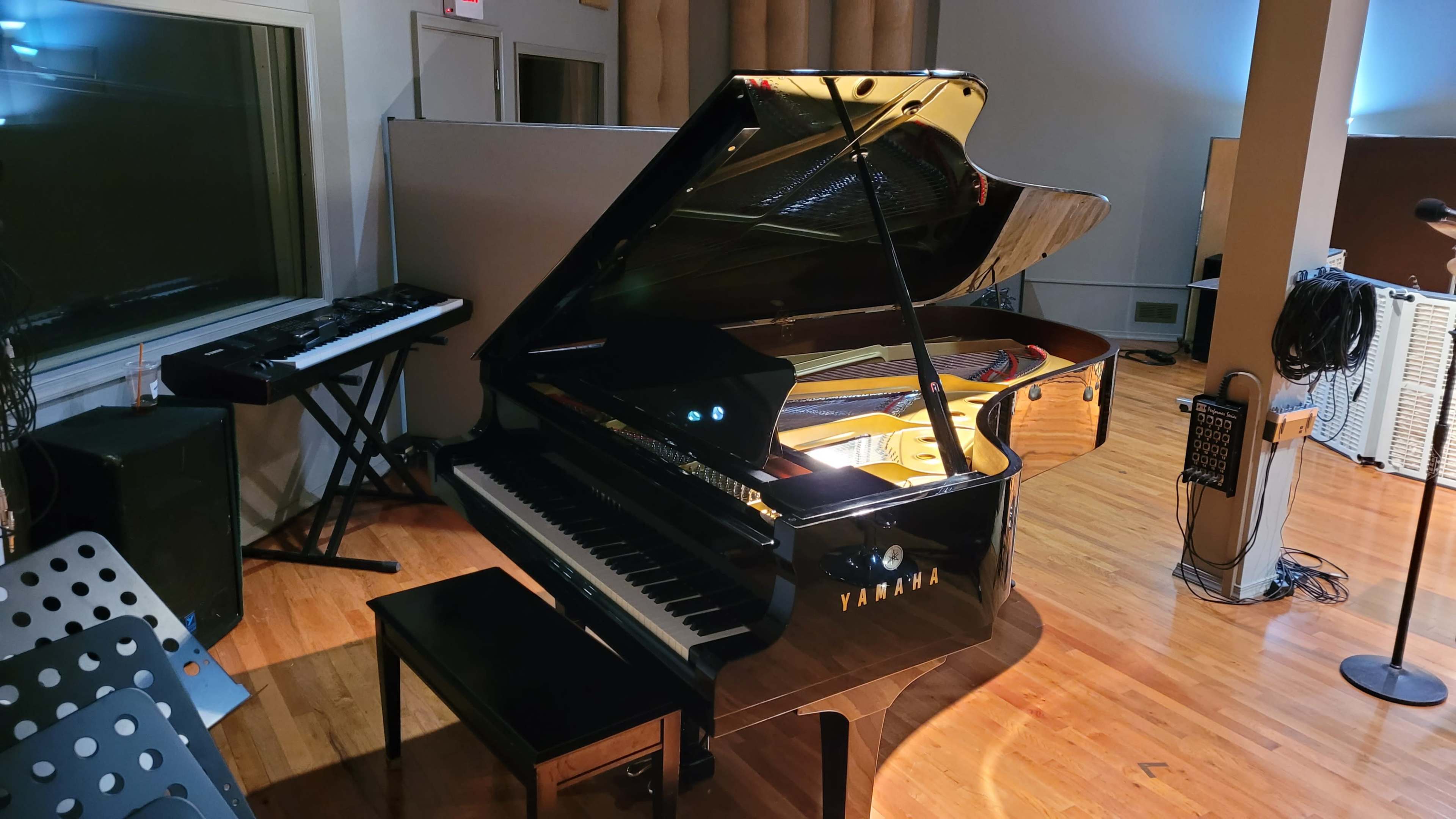 A black Yamaha grand piano is positioned in a music studio, accompanied by a keyboard and music stand in the background.