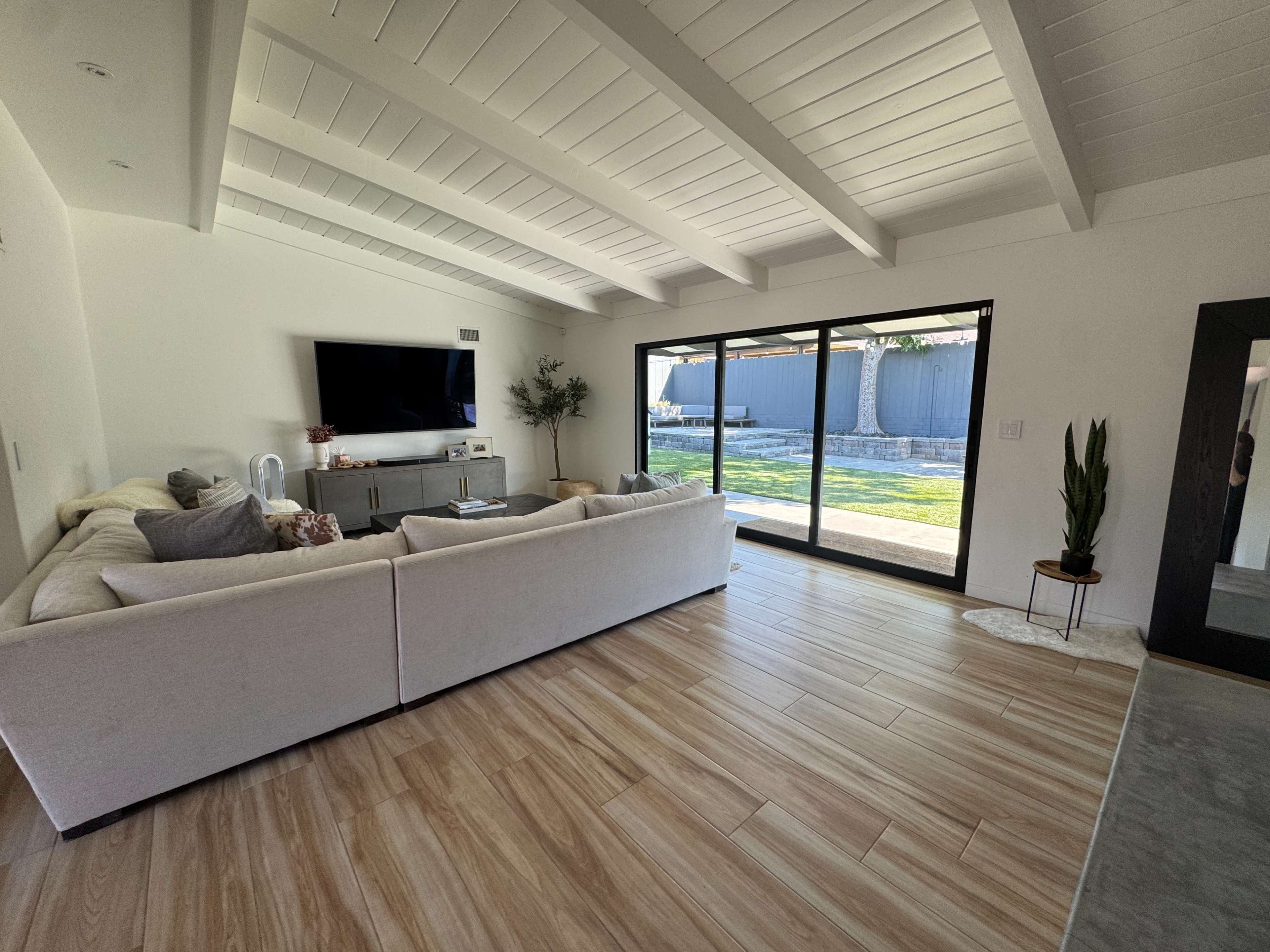 The image shows a bright living room with a large sectional sofa, a wall-mounted television, and sliding glass doors that open to a grassy outdoor area.