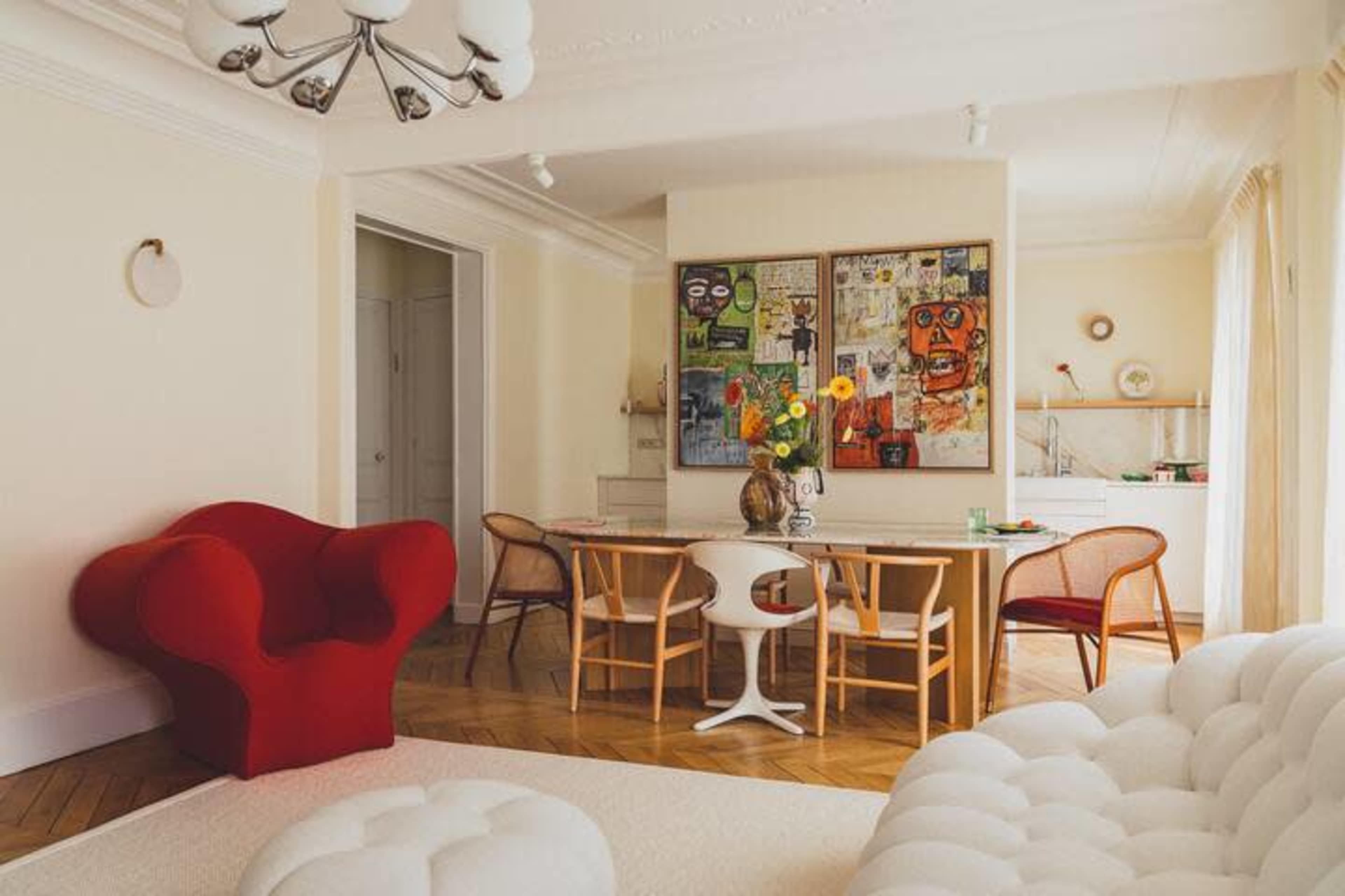 A stylish living room featuring a red chair, a wooden dining table surrounded by chairs, and colorful artwork on the wall.