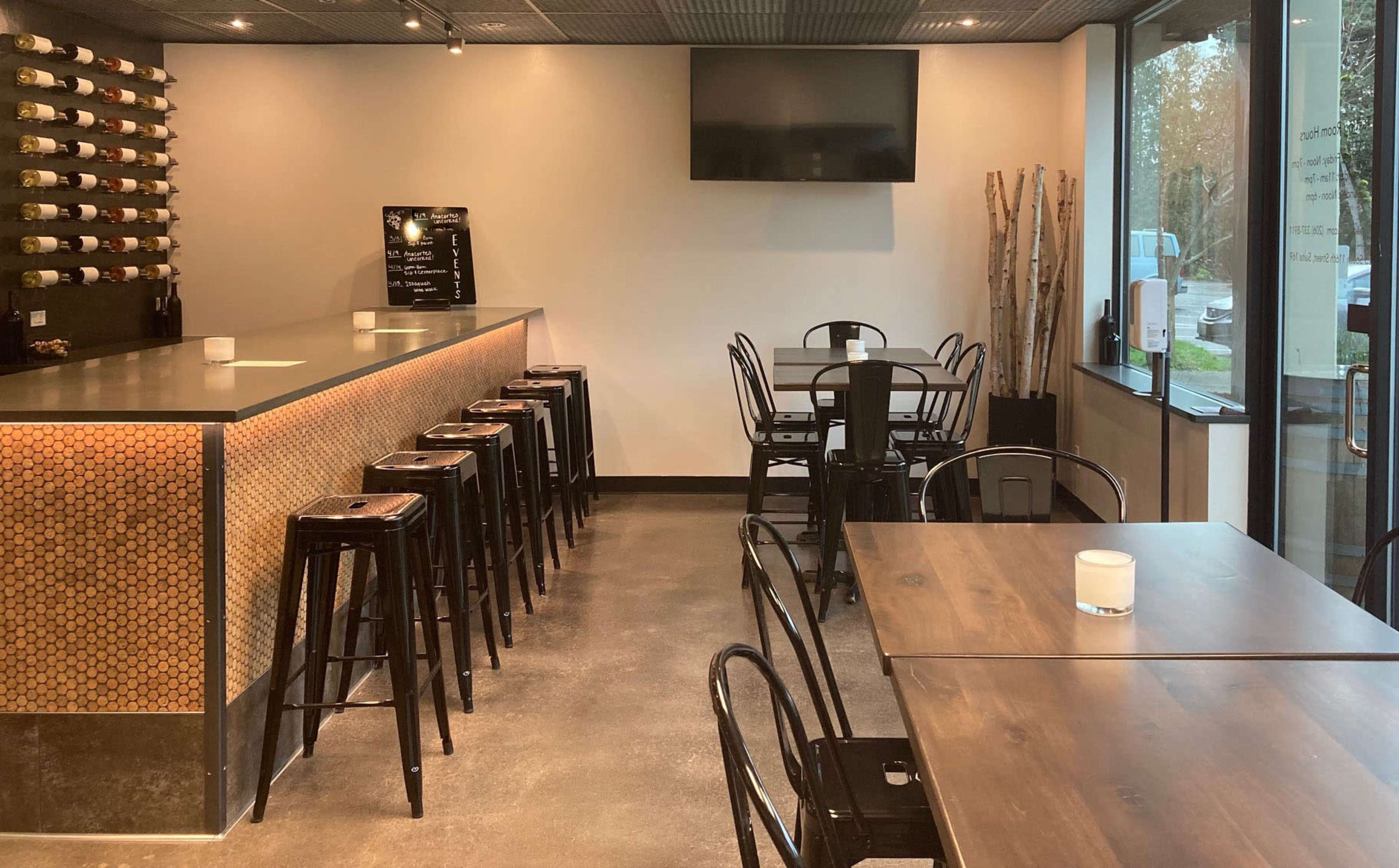 A modern wine bar featuring a long counter with high stools, scattered tables, and a wall lined with wine bottles.