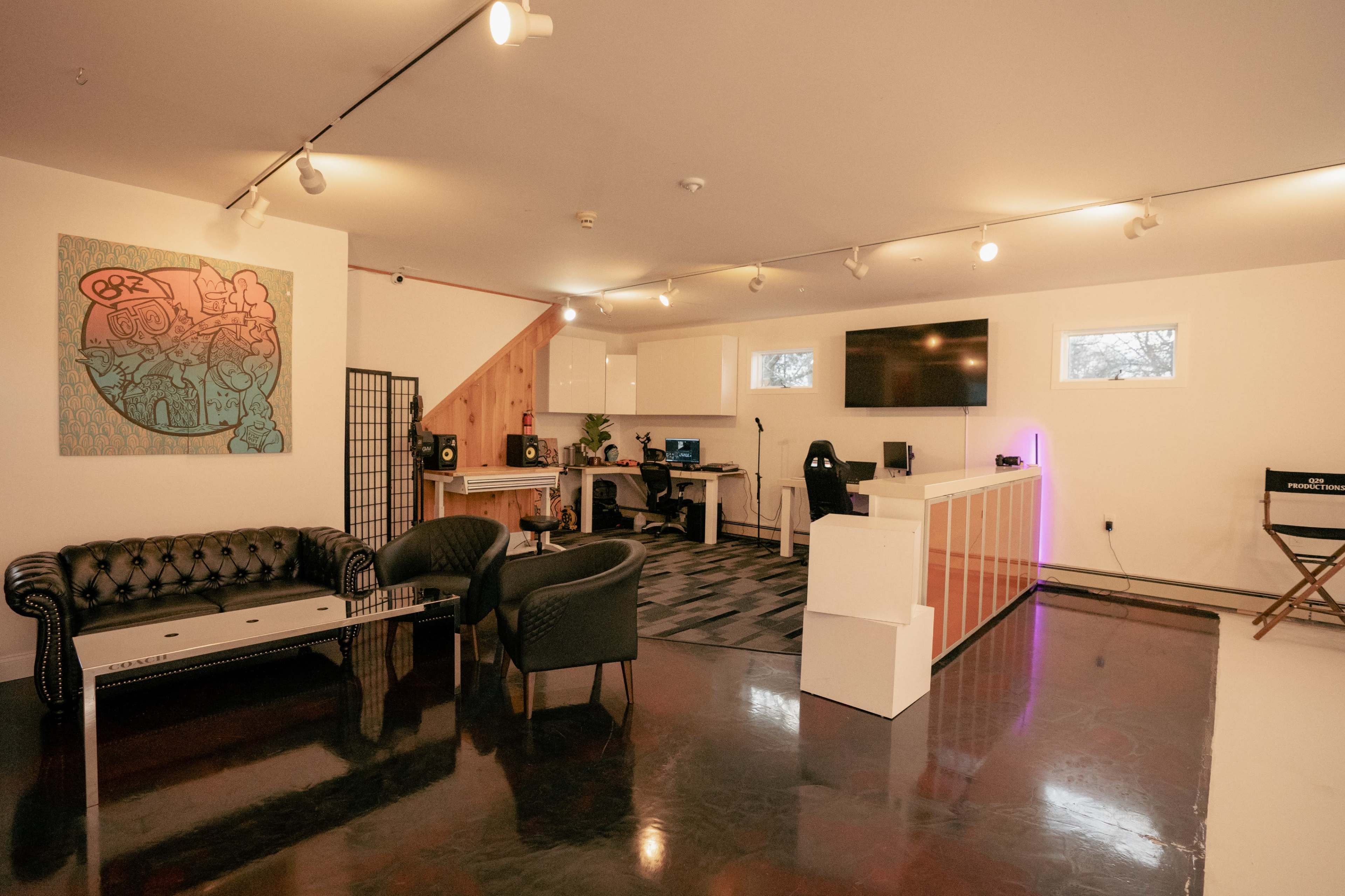 A modern, well-lit studio space with a black leather couch, two chairs, a white reception desk, and a large screen on the wall, while colorful artwork adorns one side.