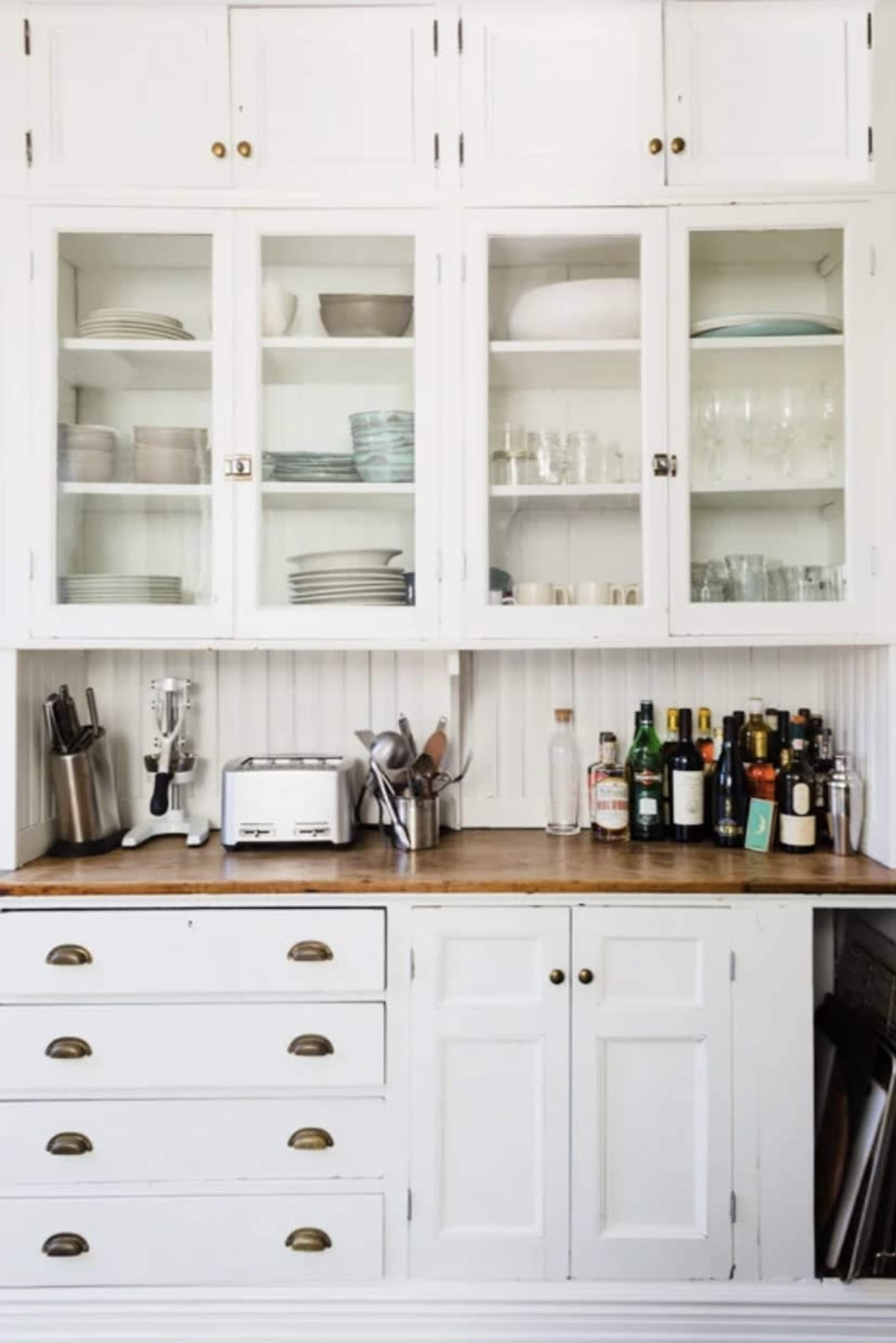 The kitchen features a white cabinetry unit with glass-fronted upper shelves displaying dishes and glasses, and a wooden countertop with various kitchen appliances and bottles organized below.