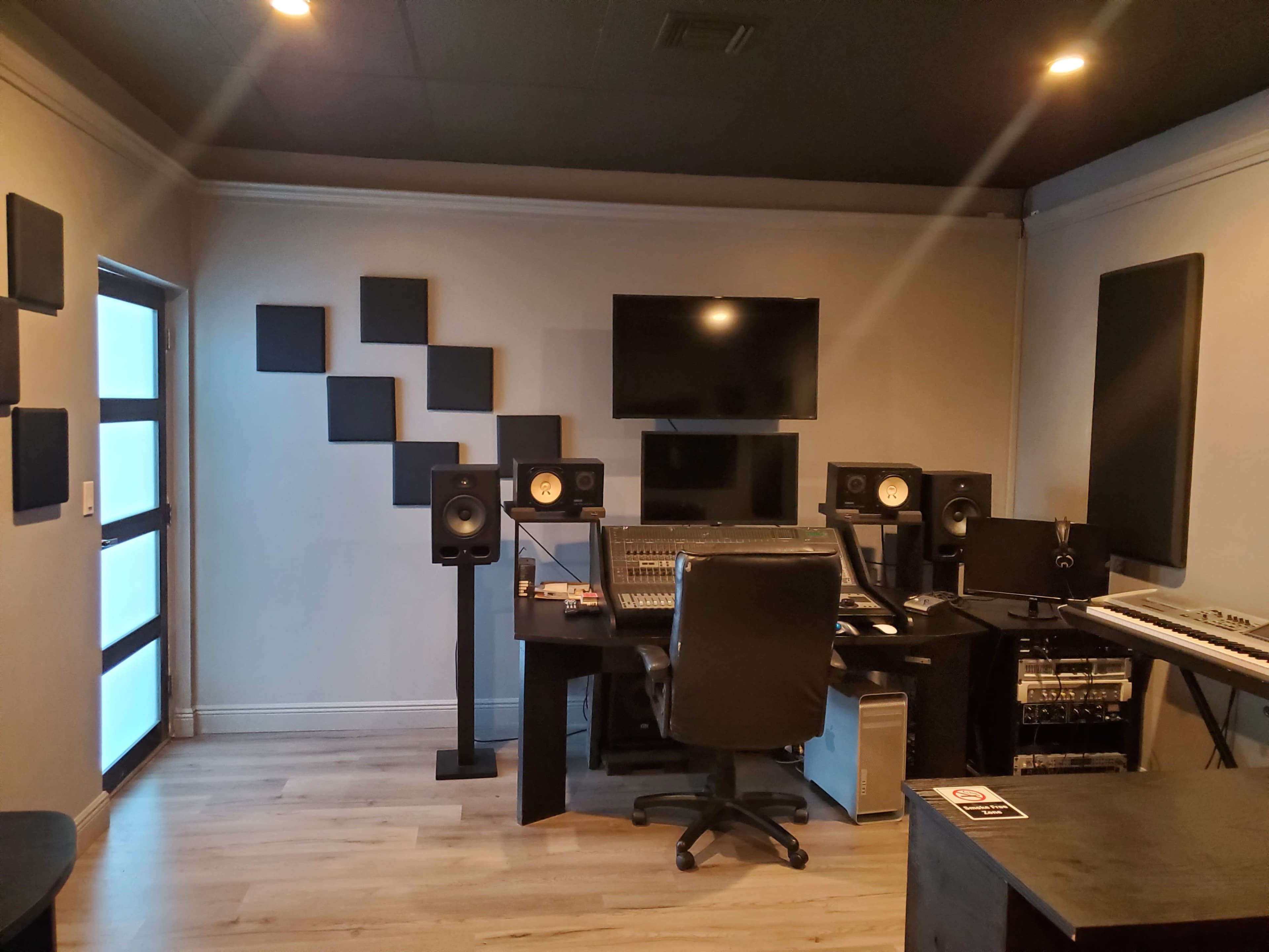 A music recording studio featuring a mixing console, two speakers on stands, a computer setup, and soundproofing panels on the walls.