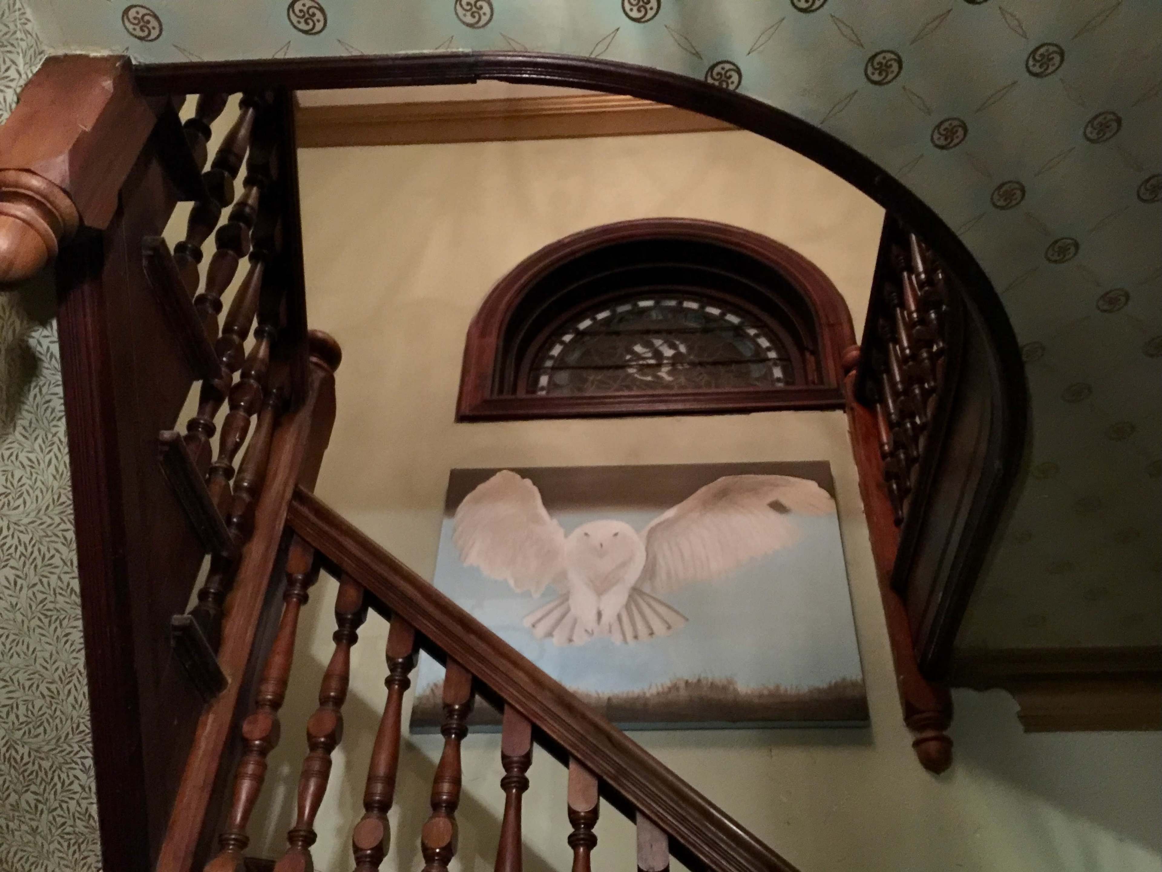 A staircase with wooden railing leads up to a landing where a large painting of an owl is mounted on the wall beneath an arched window.