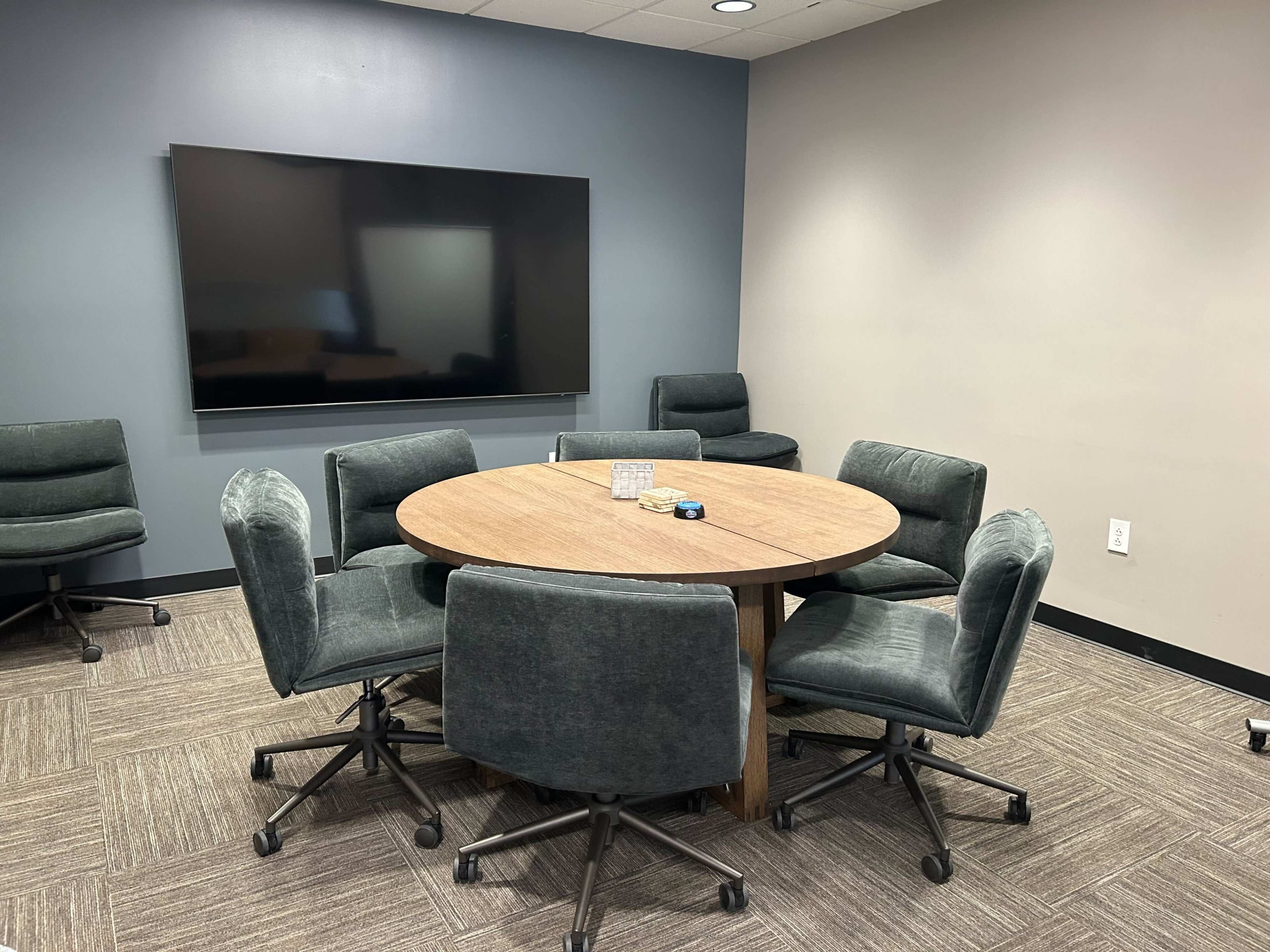 The image shows a conference room with a round wooden table surrounded by six upholstered chairs, and a large television mounted on the wall.