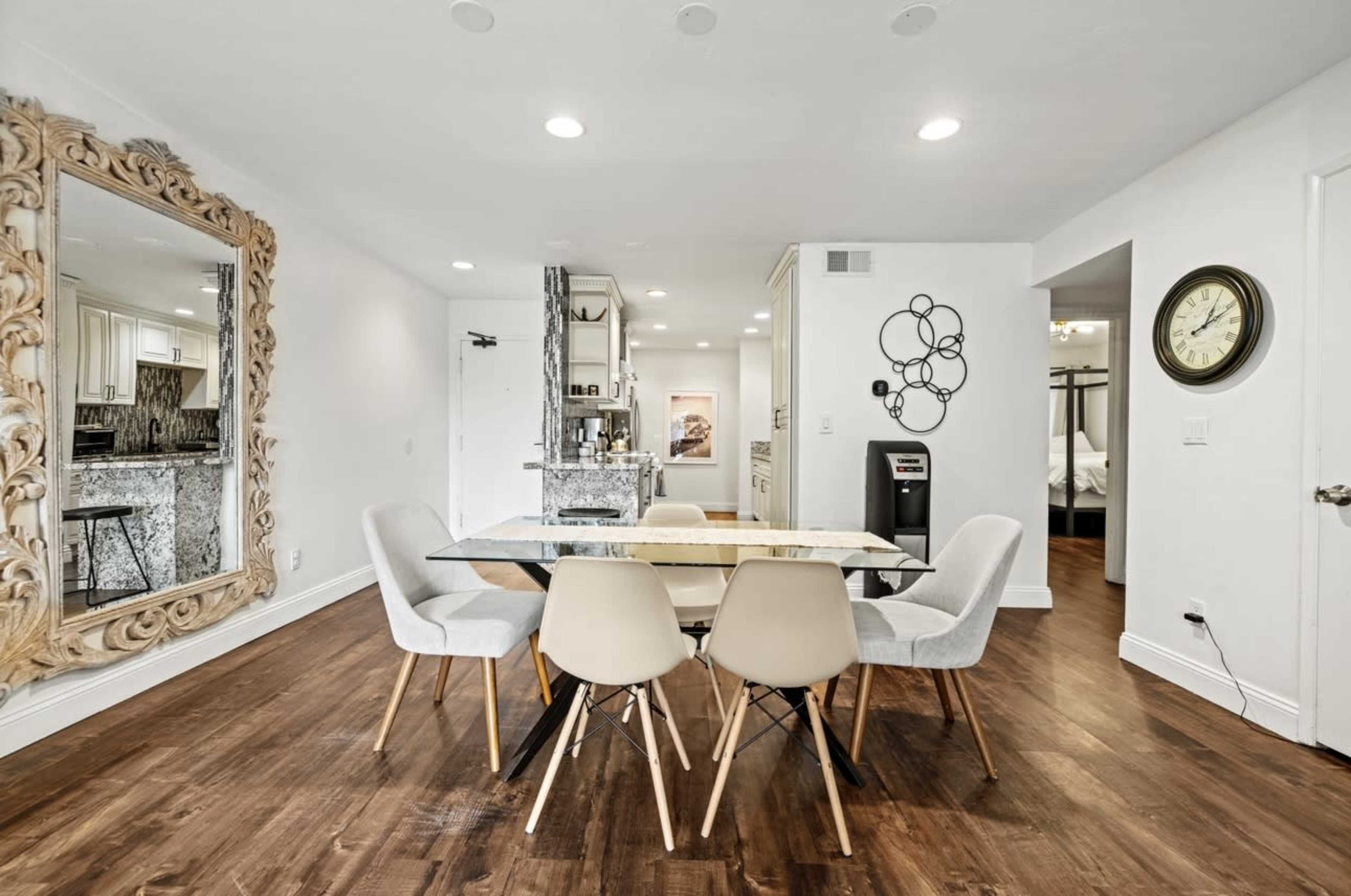 The image shows a modern dining area with a glass table surrounded by six chairs, a large decorative mirror on the wall, and a view into a kitchen with granite countertops.