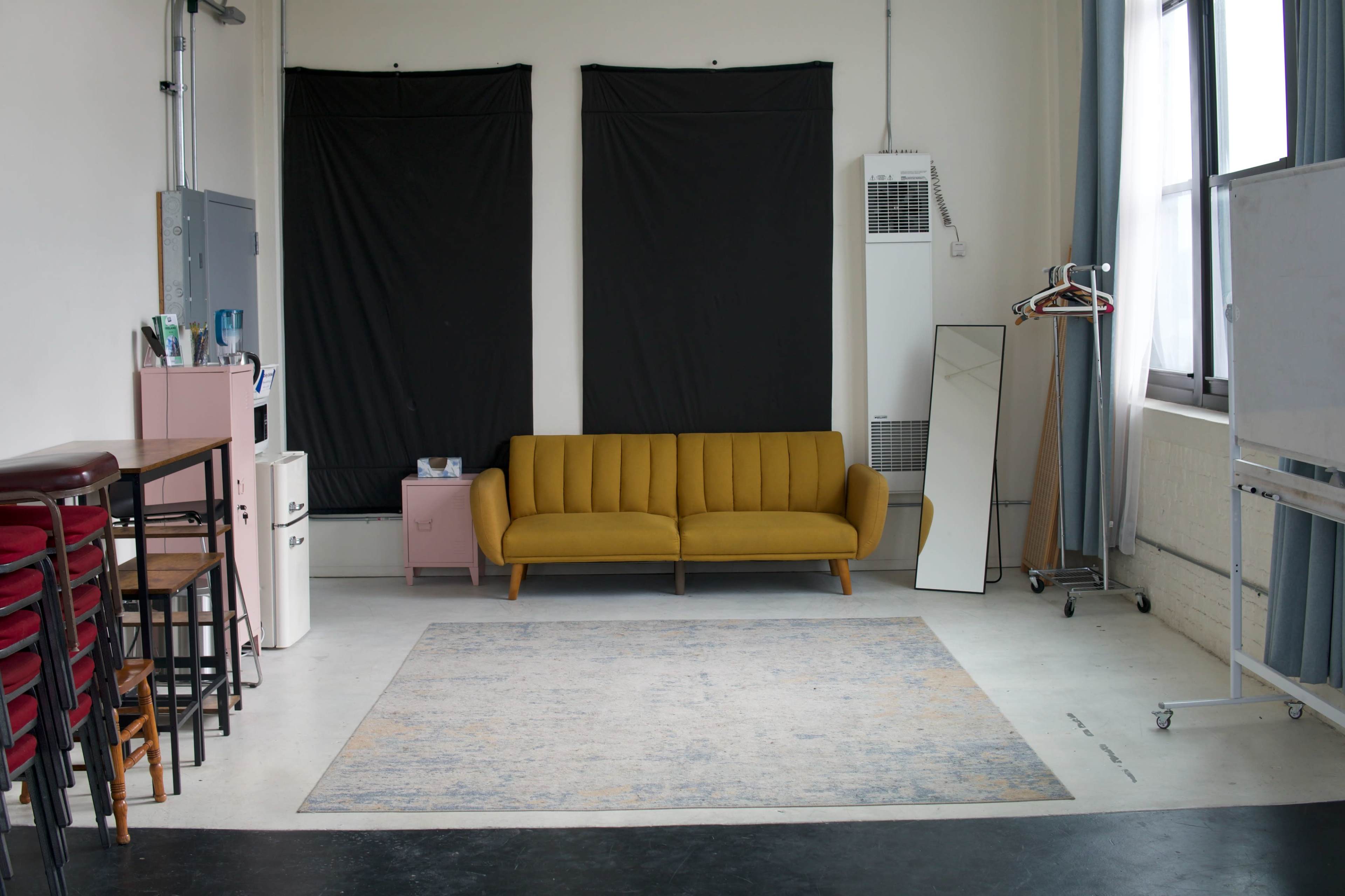 The image shows a minimalist room with a yellow sofa, a large mirror, and two black curtains covering windows, along with a small pink nightstand and some furniture against the walls.