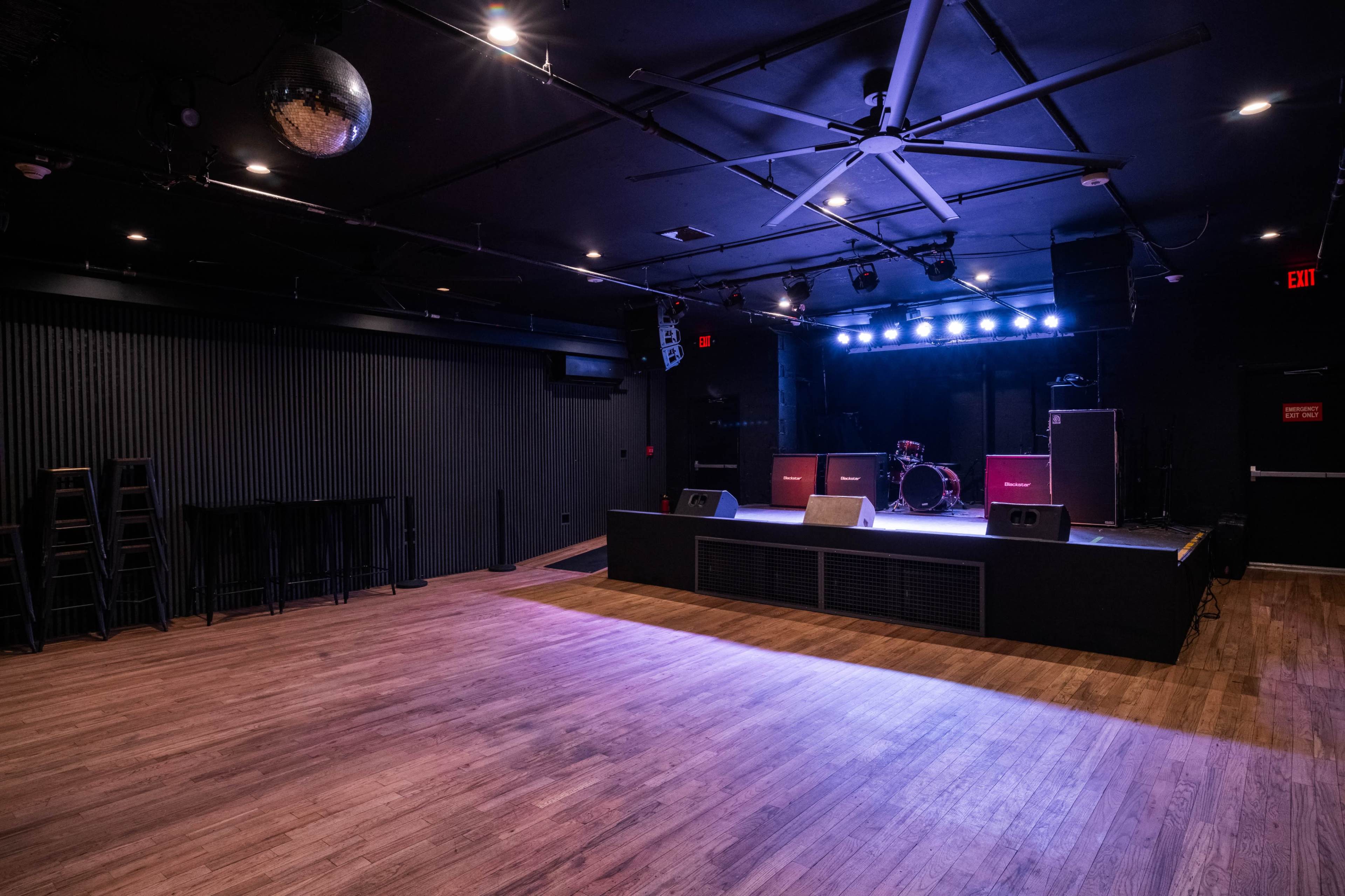 The image shows a dimly lit stage area in a music venue with a wooden floor, a drum set, amplifiers, and stage lights.