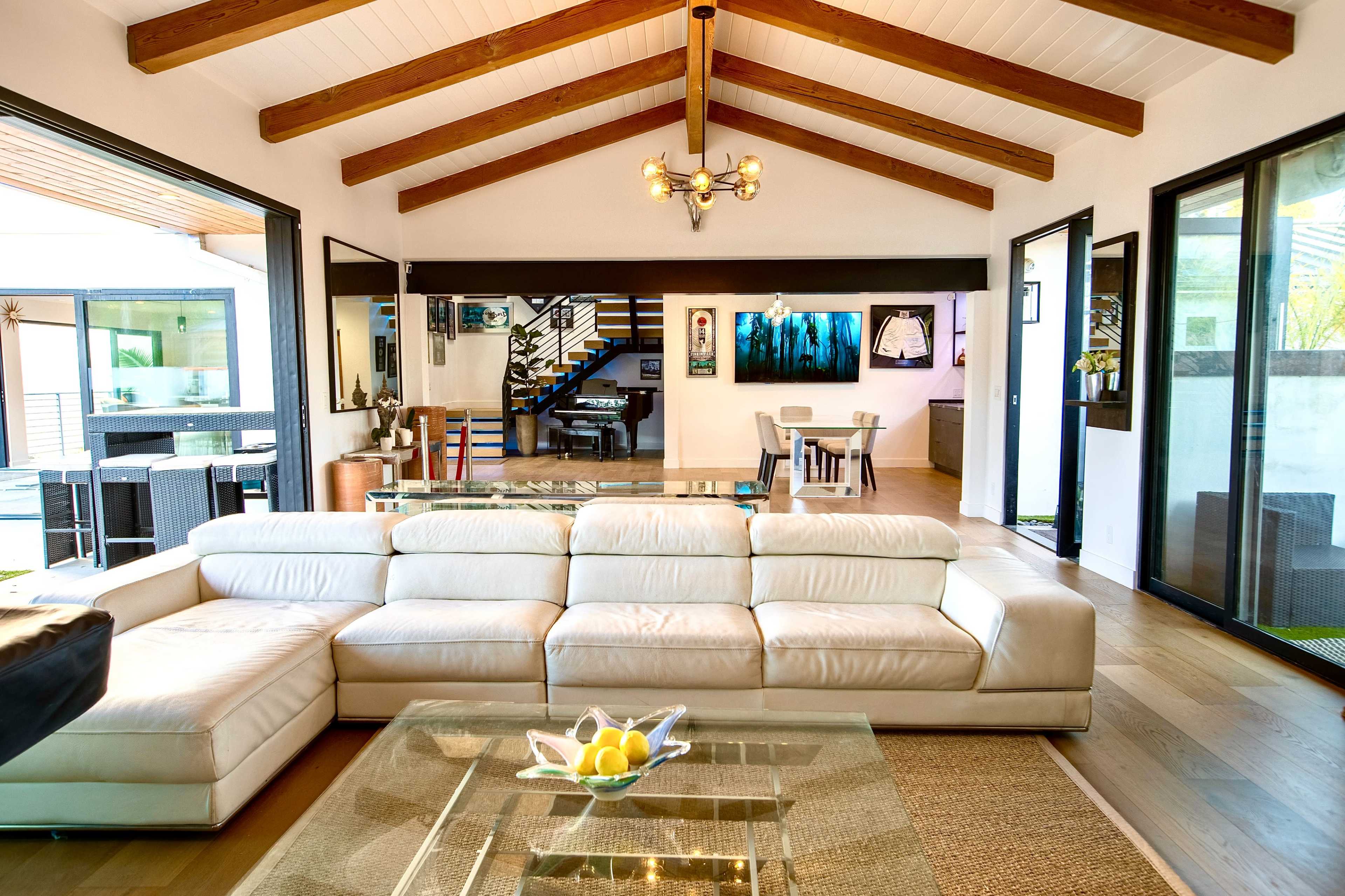 The image shows a spacious living room with a white leather sectional sofa, wooden beam ceilings, and large glass doors leading to an outdoor area.