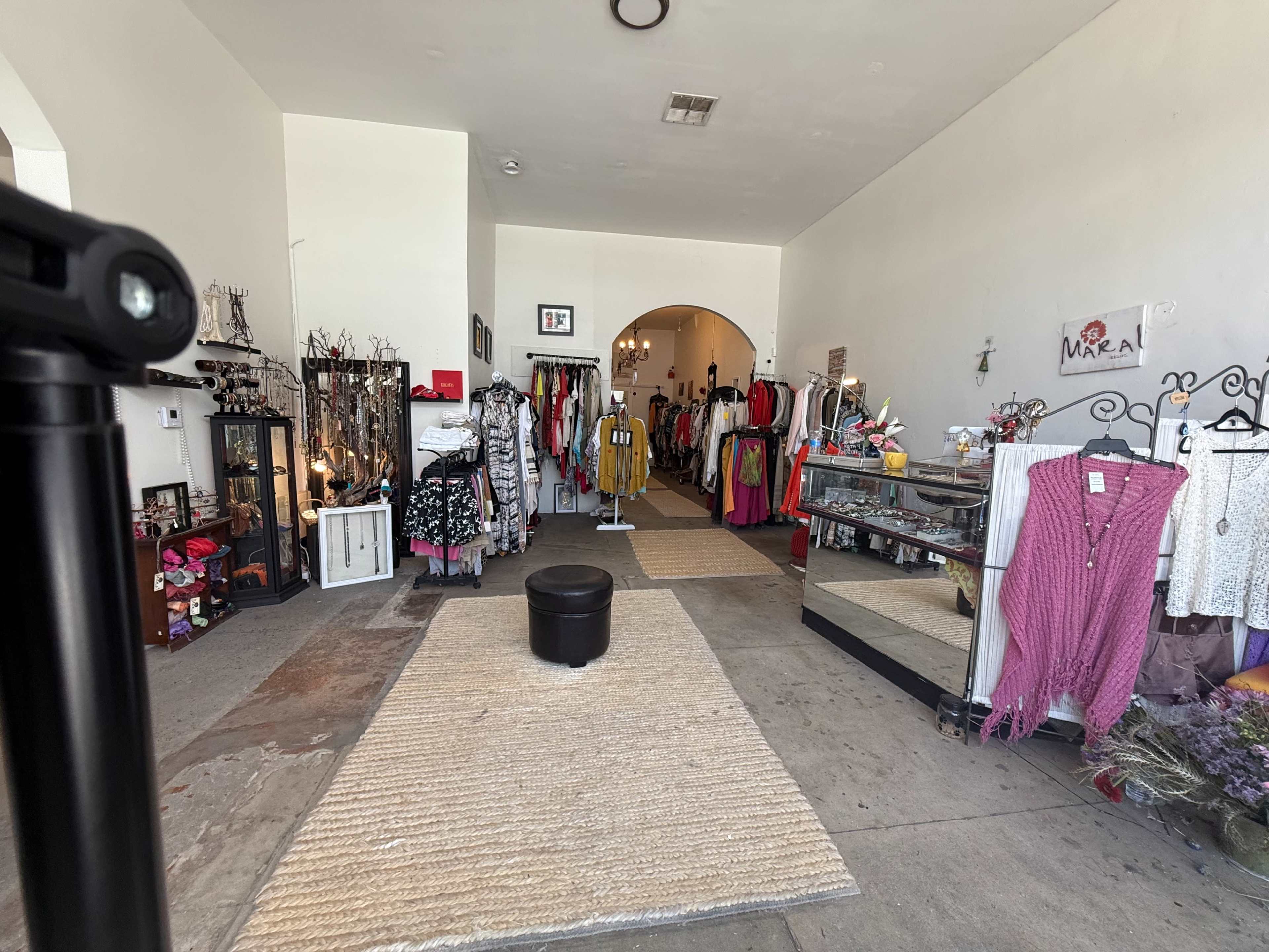 The image shows the interior of a clothing store displaying various garments, accessories, and a central seating area.