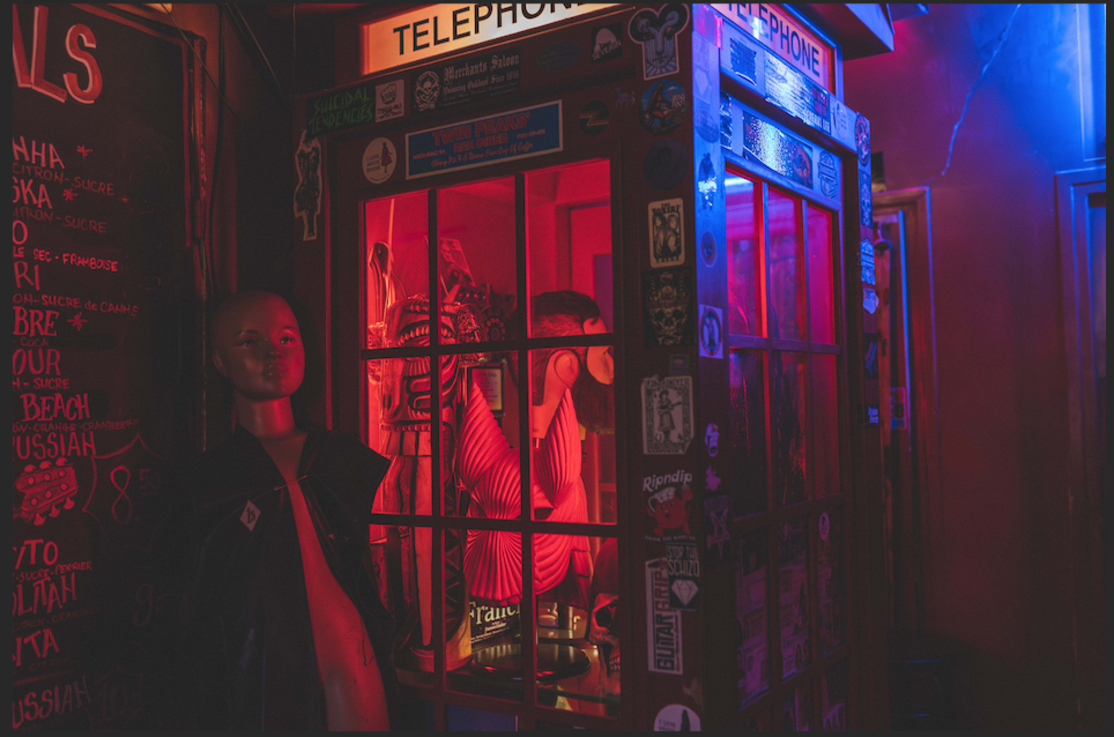 A brightly lit vintage telephone booth adorned with stickers stands next to a mannequin in a dimly lit space.