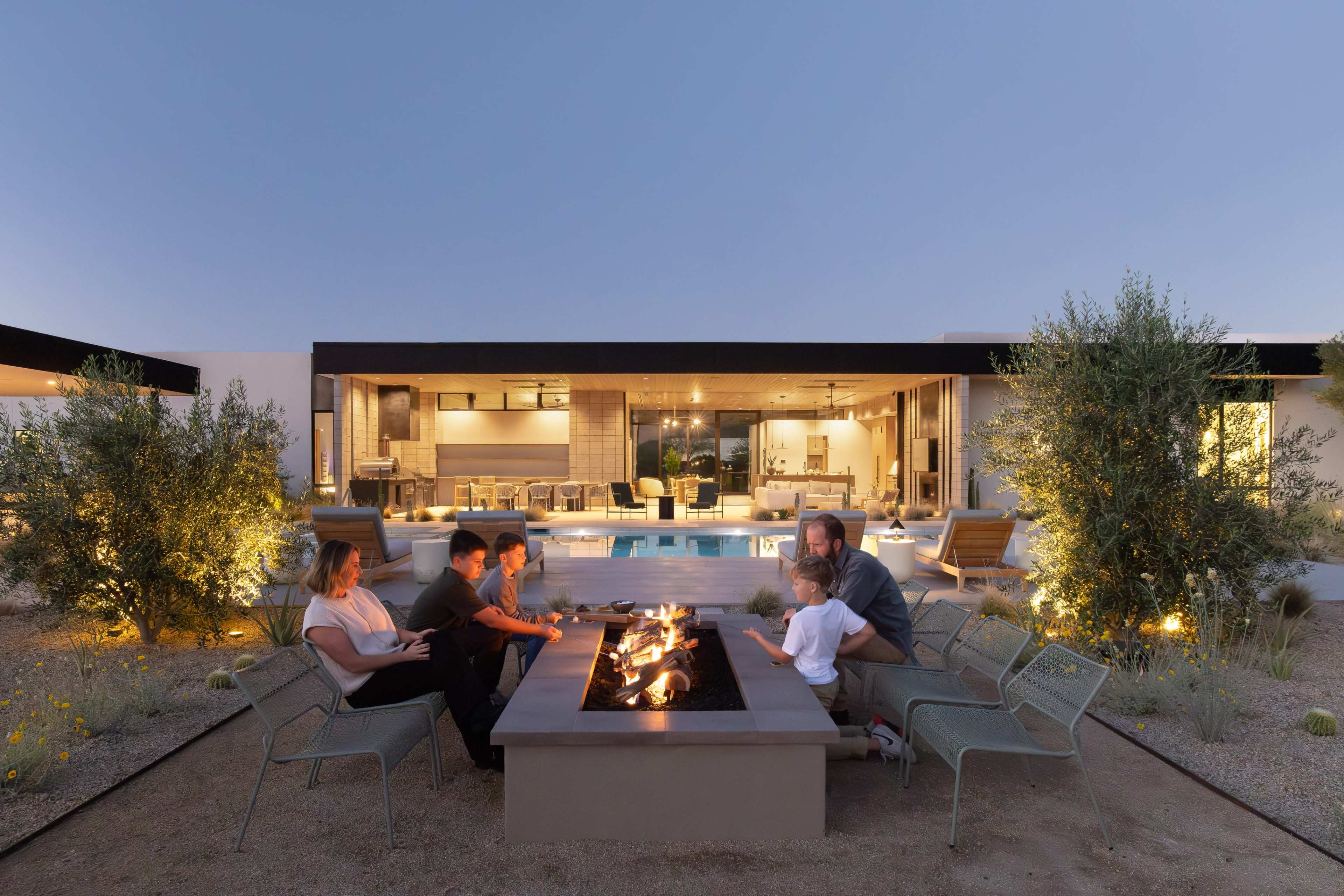 A family sits around a fire pit in an outdoor setting, with a modern home and swimming pool visible in the background.