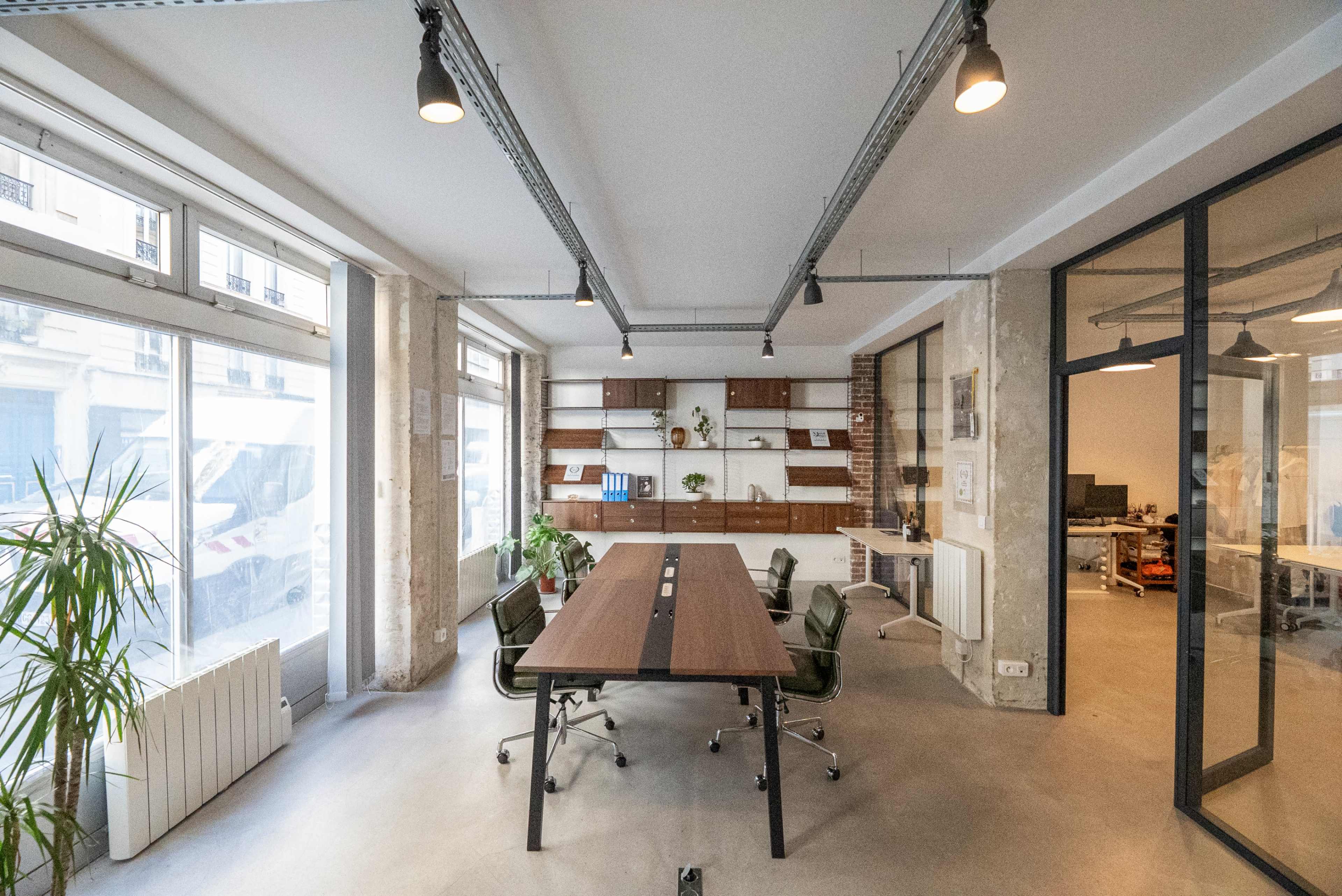 The image shows a modern office space featuring a large wooden conference table surrounded by chairs, with shelves displaying plants and books in the background.