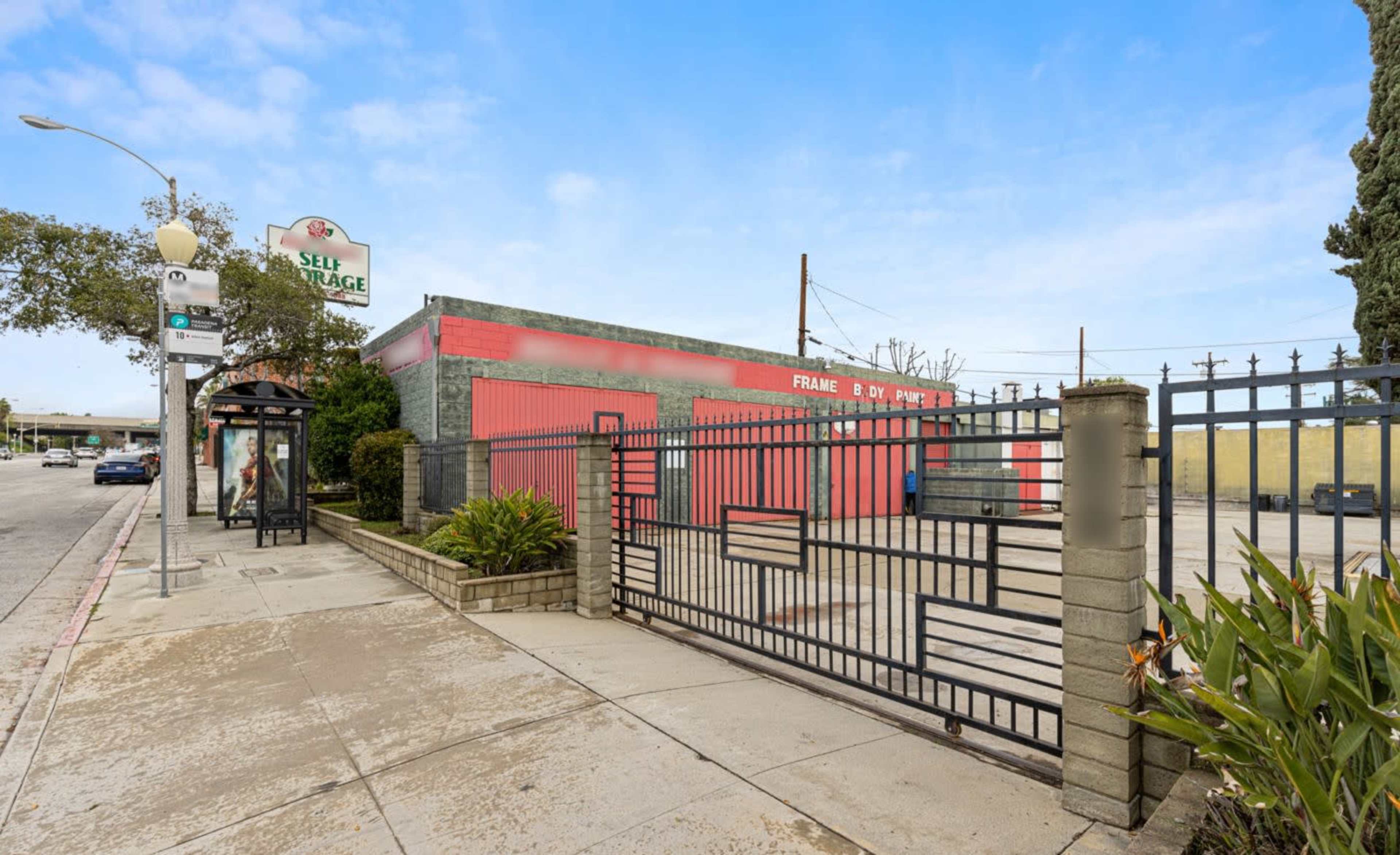 A self-storage facility with a red exterior and a gated entrance, located on a city street next to a bus stop.