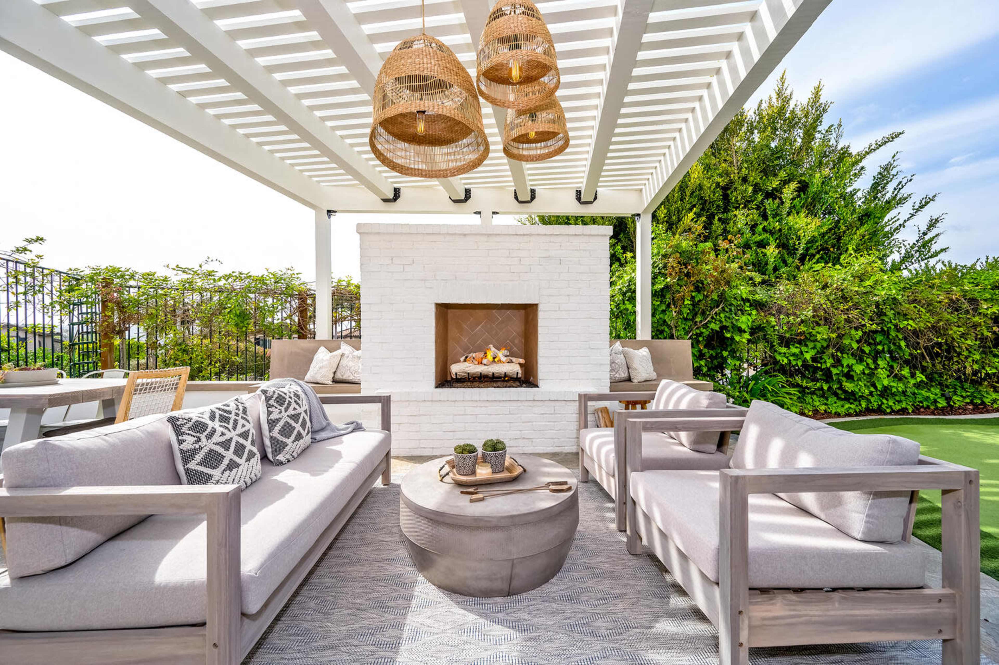 A stylish outdoor lounge area with two gray sofas, a round coffee table, and a white brick fireplace, all set under a white pergola with hanging light fixtures.