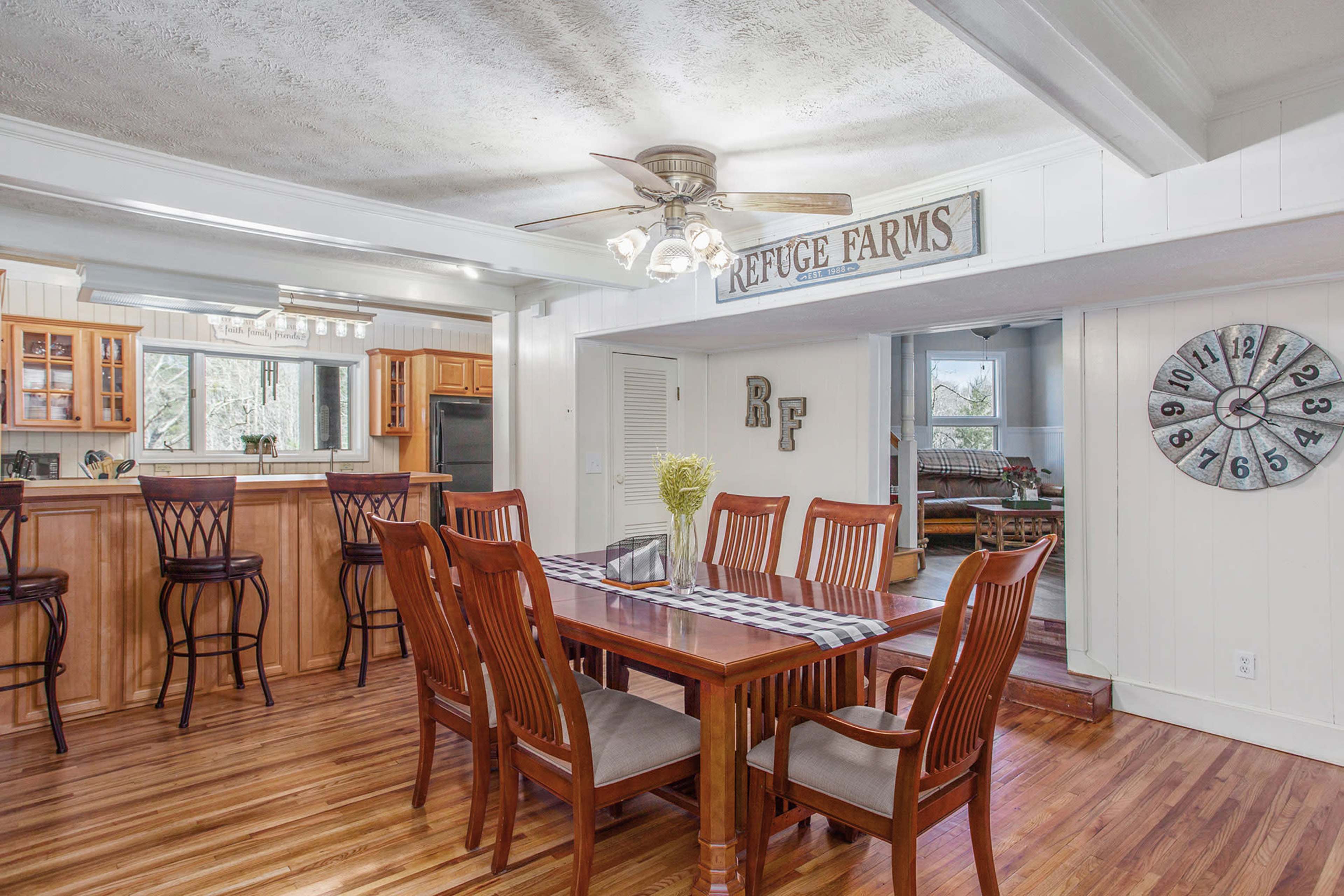 The image shows a spacious dining area with a wooden table and chairs, a ceiling fan, and a view into a kitchen and living area.