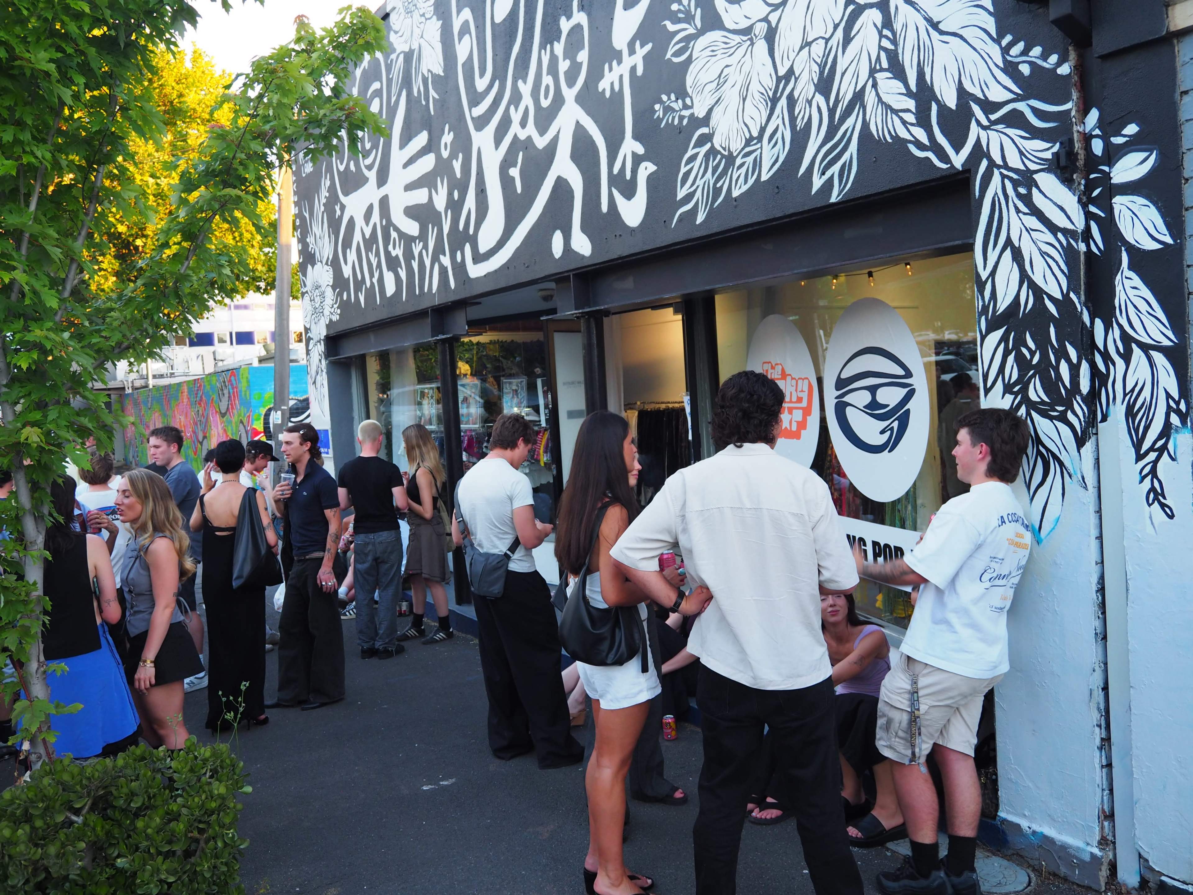 A crowd gathers outside a shop adorned with colorful murals, engaging in conversation during an event.