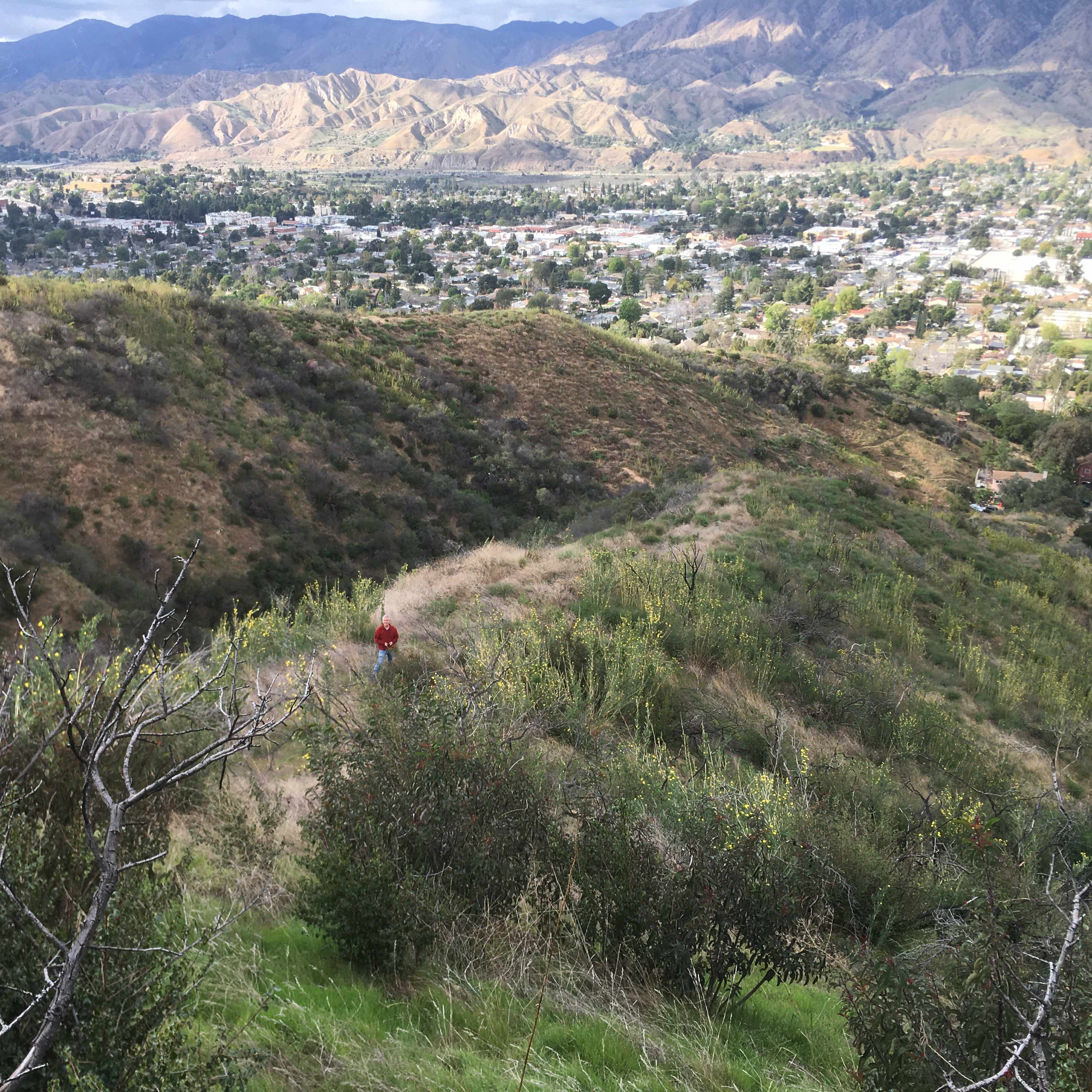 A lone figure walks along a hillside trail overlooking a valley and distant mountains.