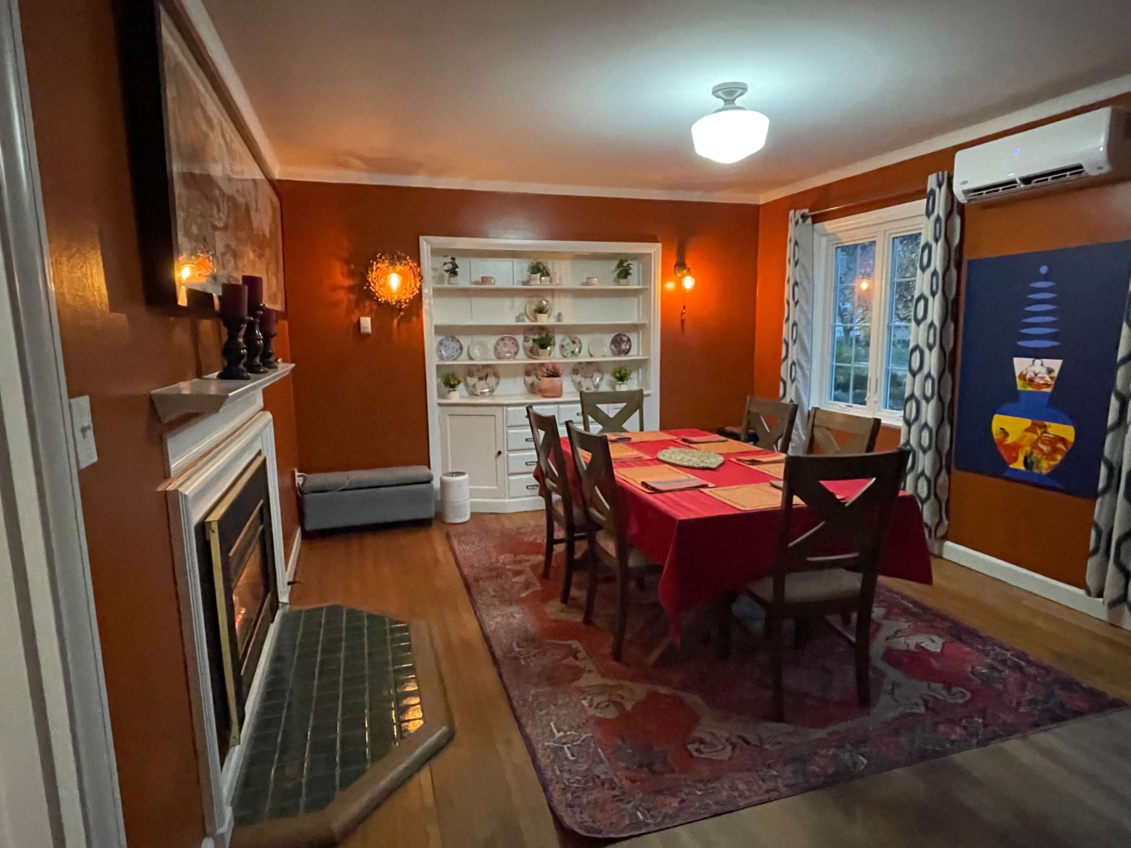 A dining room features a red tablecloth over a large table surrounded by chairs, with a fireplace and shelves displaying dishes along the walls.