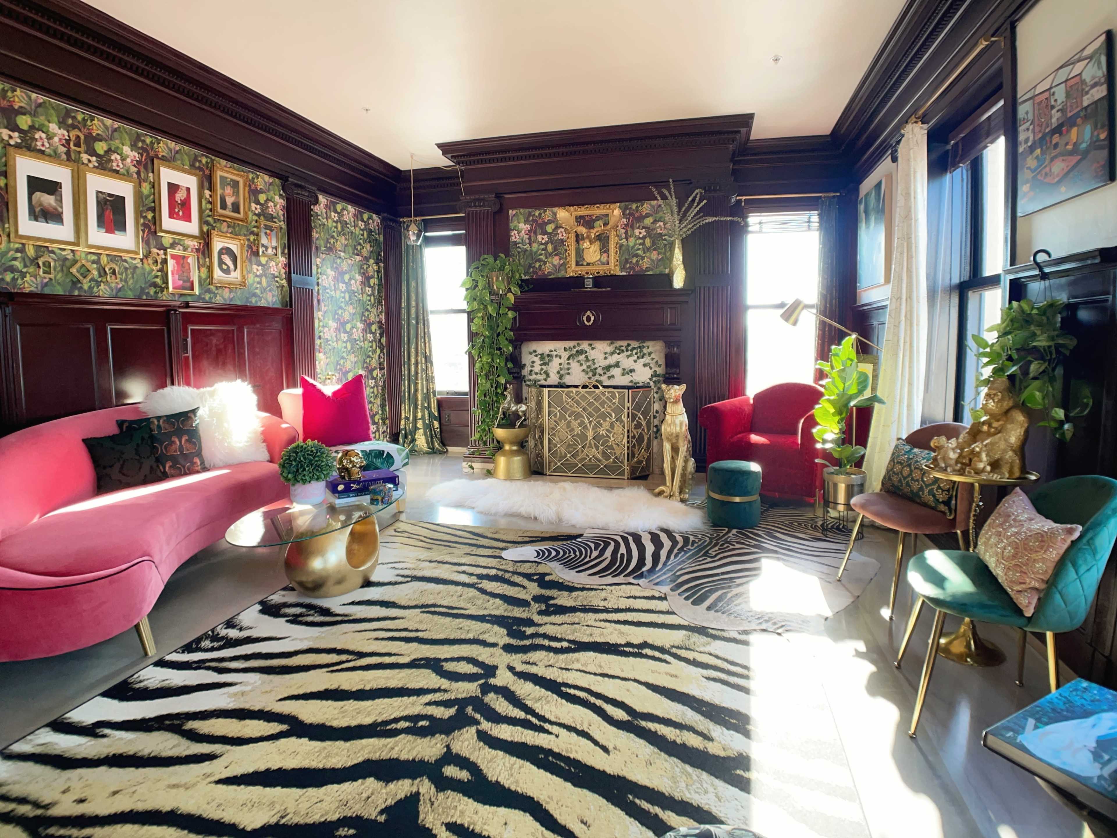 A vibrant living room with a pink sofa, red armchairs, a patterned rug, and various decorative plants and artwork.