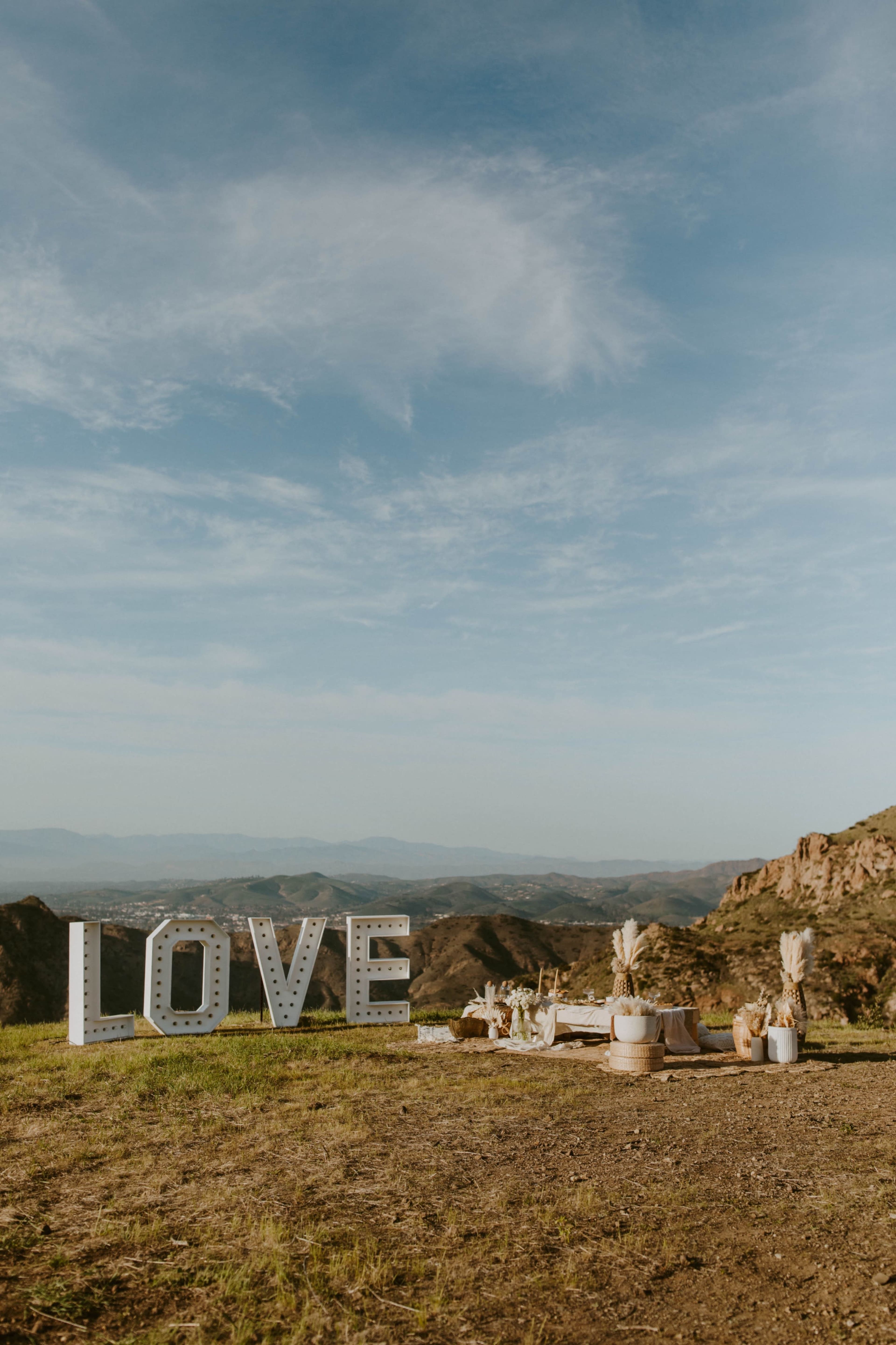 A large illuminated sign spelling "LOVE" stands on a grassy area overlooking a mountainous landscape with a picnic setup nearby.