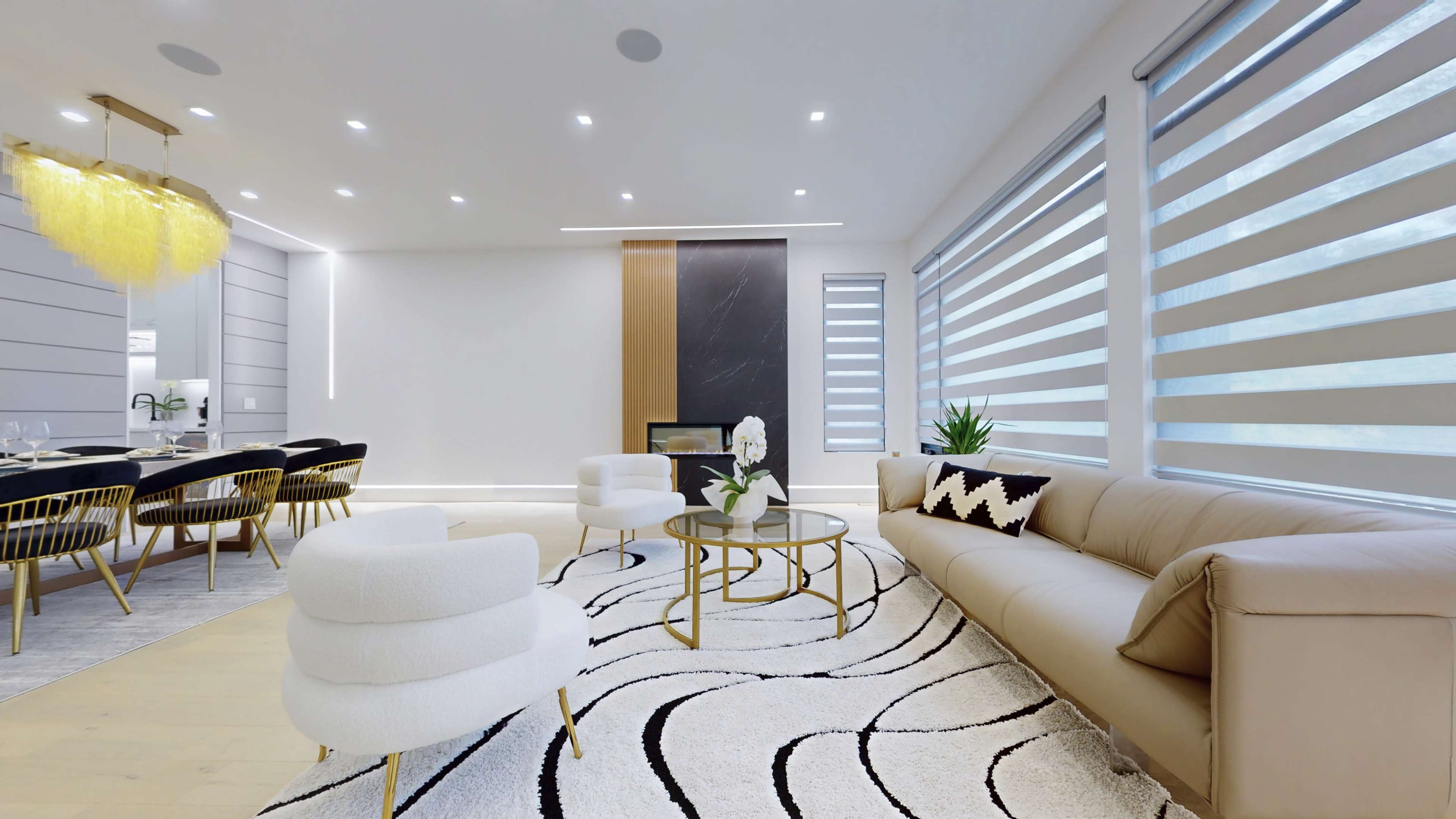 A modern living room and dining area featuring a beige sofa, a round coffee table on a patterned rug, large windows with striped shades, and a dark fireplace backdrop.