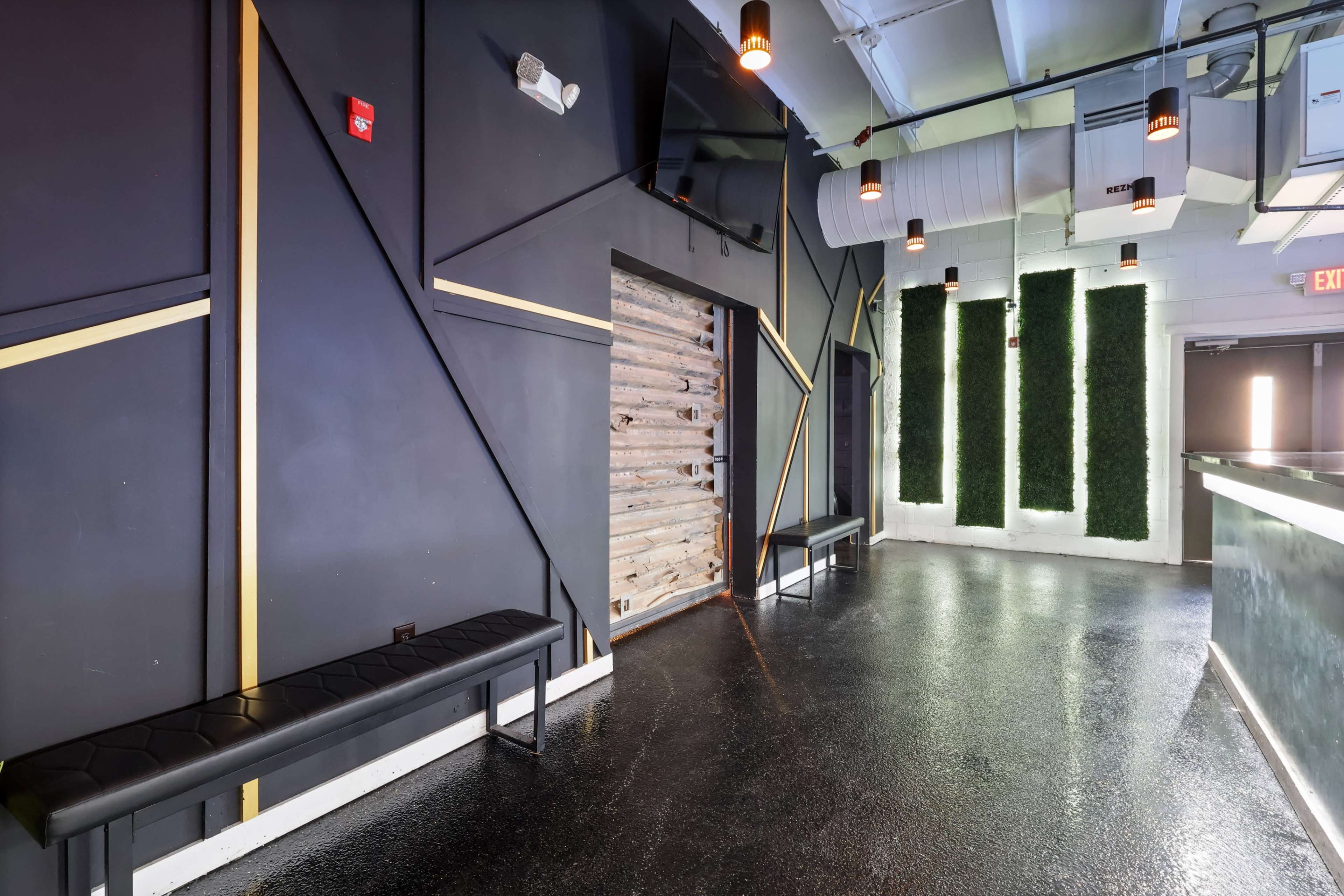The image shows an interior space with black walls, a wooden door, benches along the sides, and a bar counter at one end, featuring green vertical wall accents.