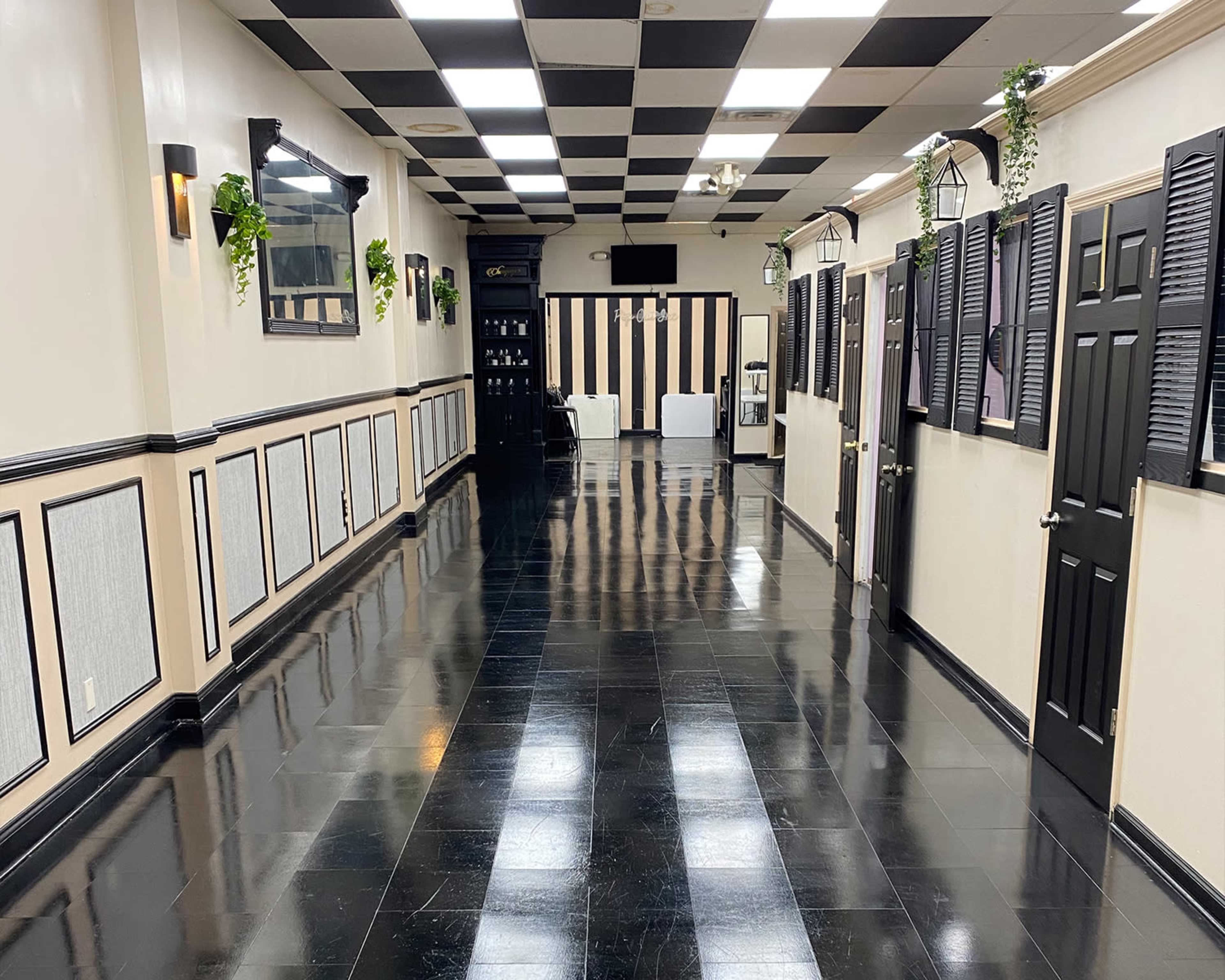 A long, narrow hallway with black and white checkered tiles, flanked by doors and wall decorations, leading to a seating area at the far end.