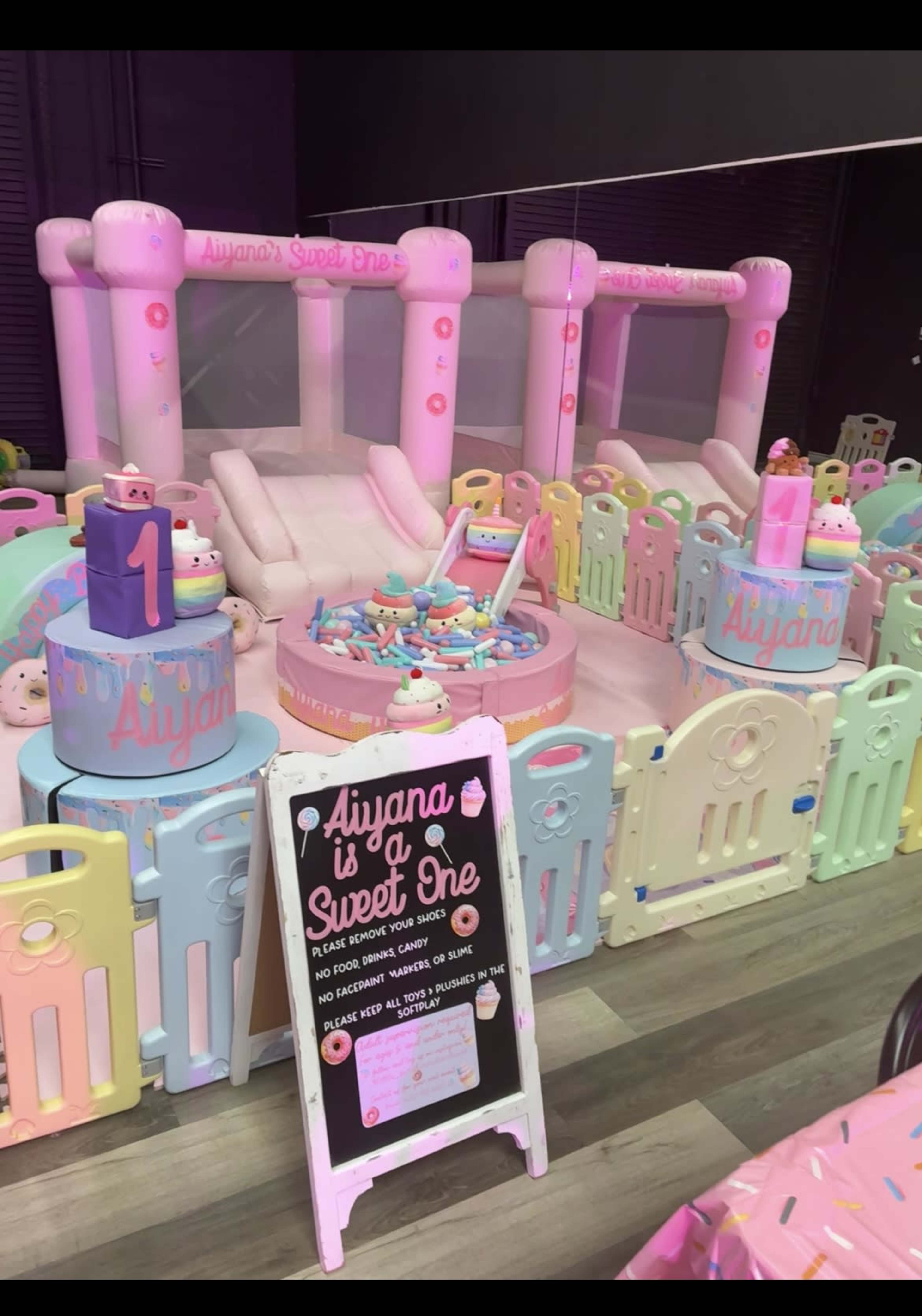 The image shows a colorful indoor children's birthday party setting with a bouncy castle, decorated tables, and a sign announcing the celebration of "Aiyana's Sweet One."