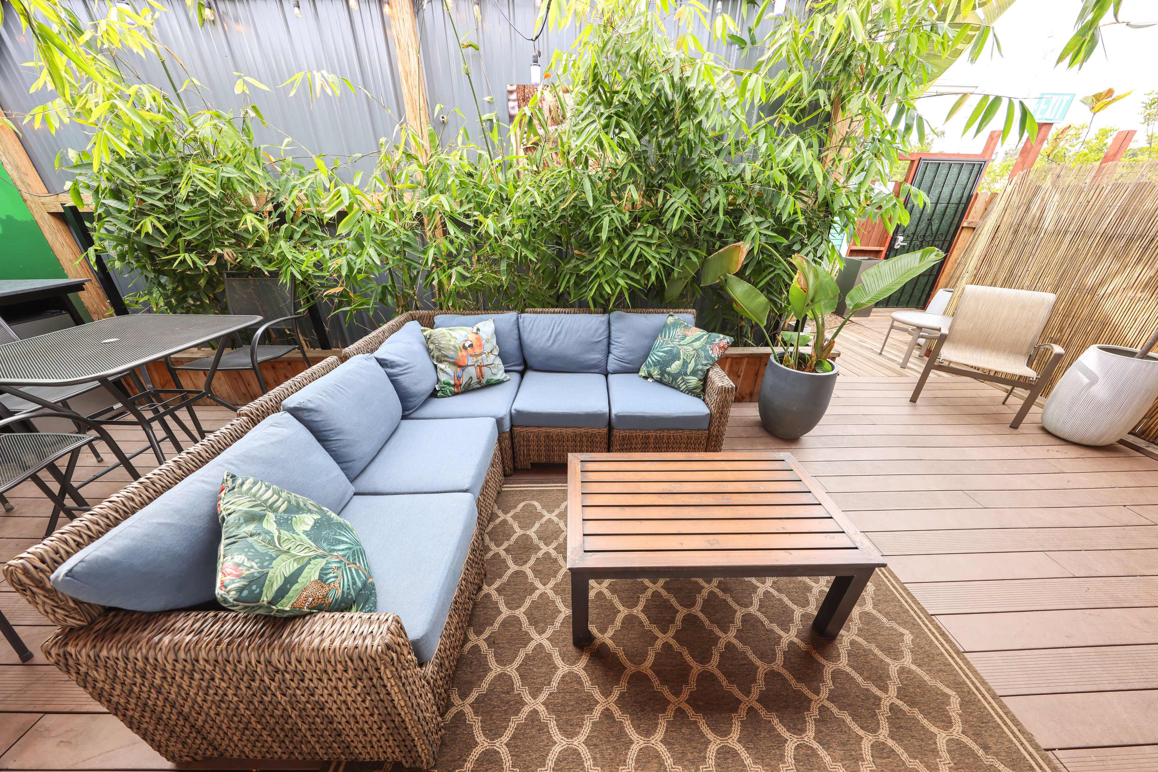 The image shows a patio area featuring a large sectional wicker couch with blue cushions, a wooden coffee table, and plants in the background.