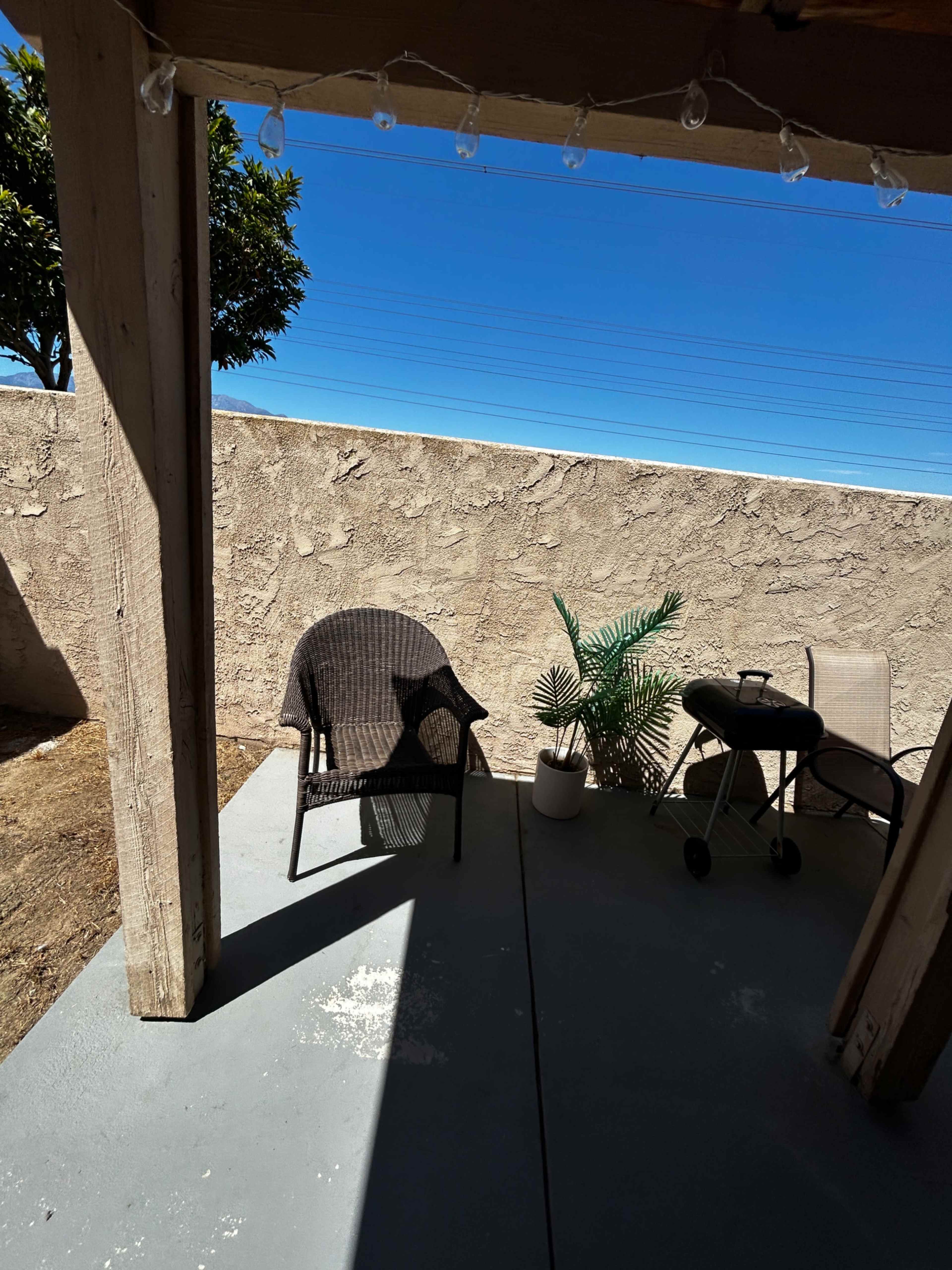 The image shows a view of a shaded patio with two chairs, a small table, and a potted plant, framed by a textured wall and a clear blue sky.