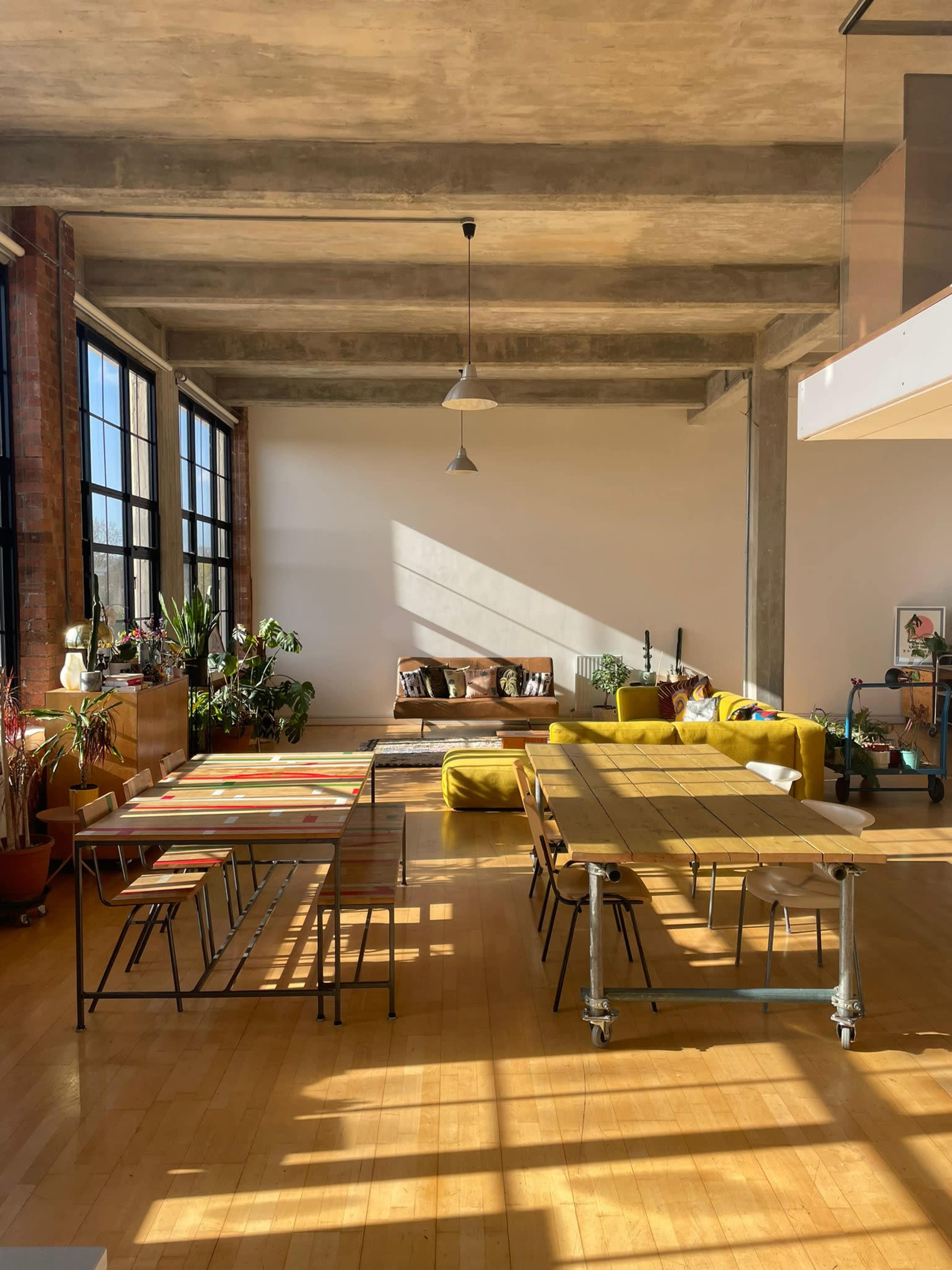 A spacious, bright room featuring large windows, a yellow couch, a dining table with metal legs, and various plants.