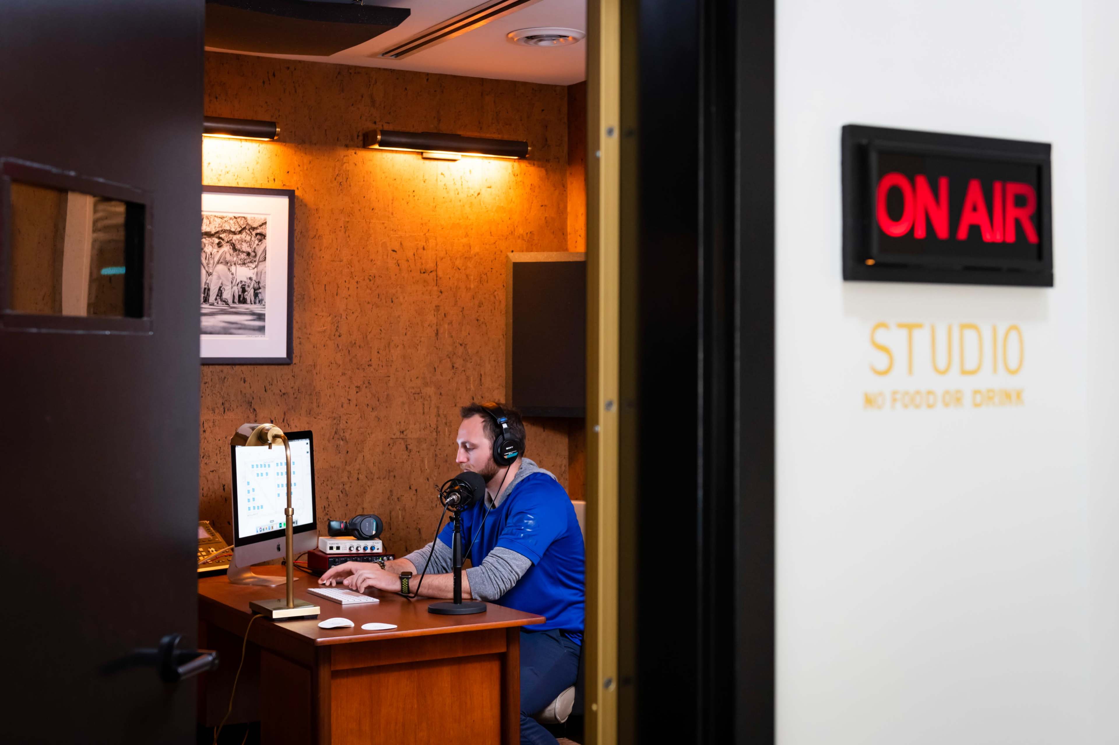 A person wearing headphones sits at a desk with a microphone and computer in a recording studio marked "ON AIR."