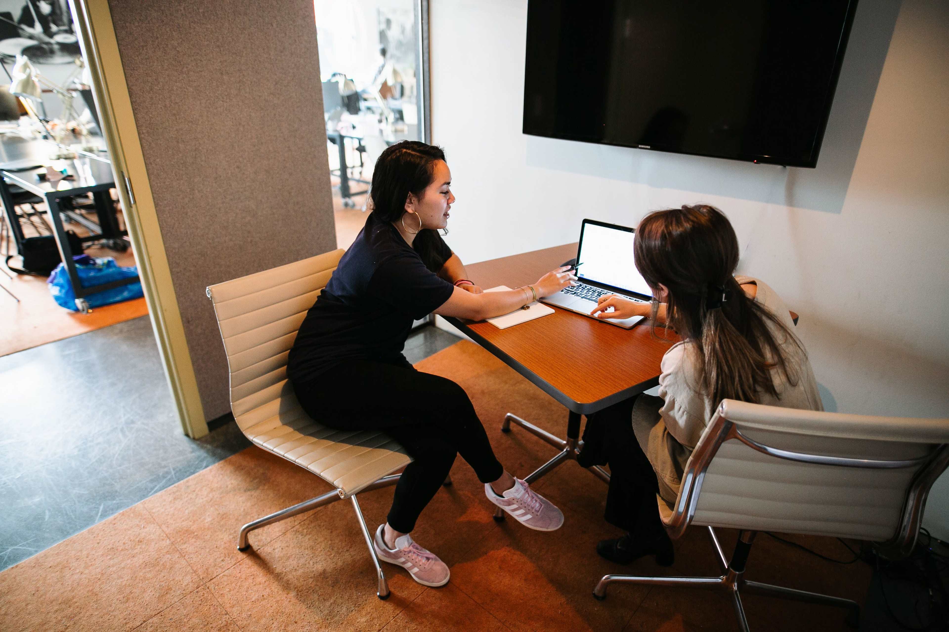 Two women sit at a table in a meeting room, one pointing at a laptop screen while the other takes notes.