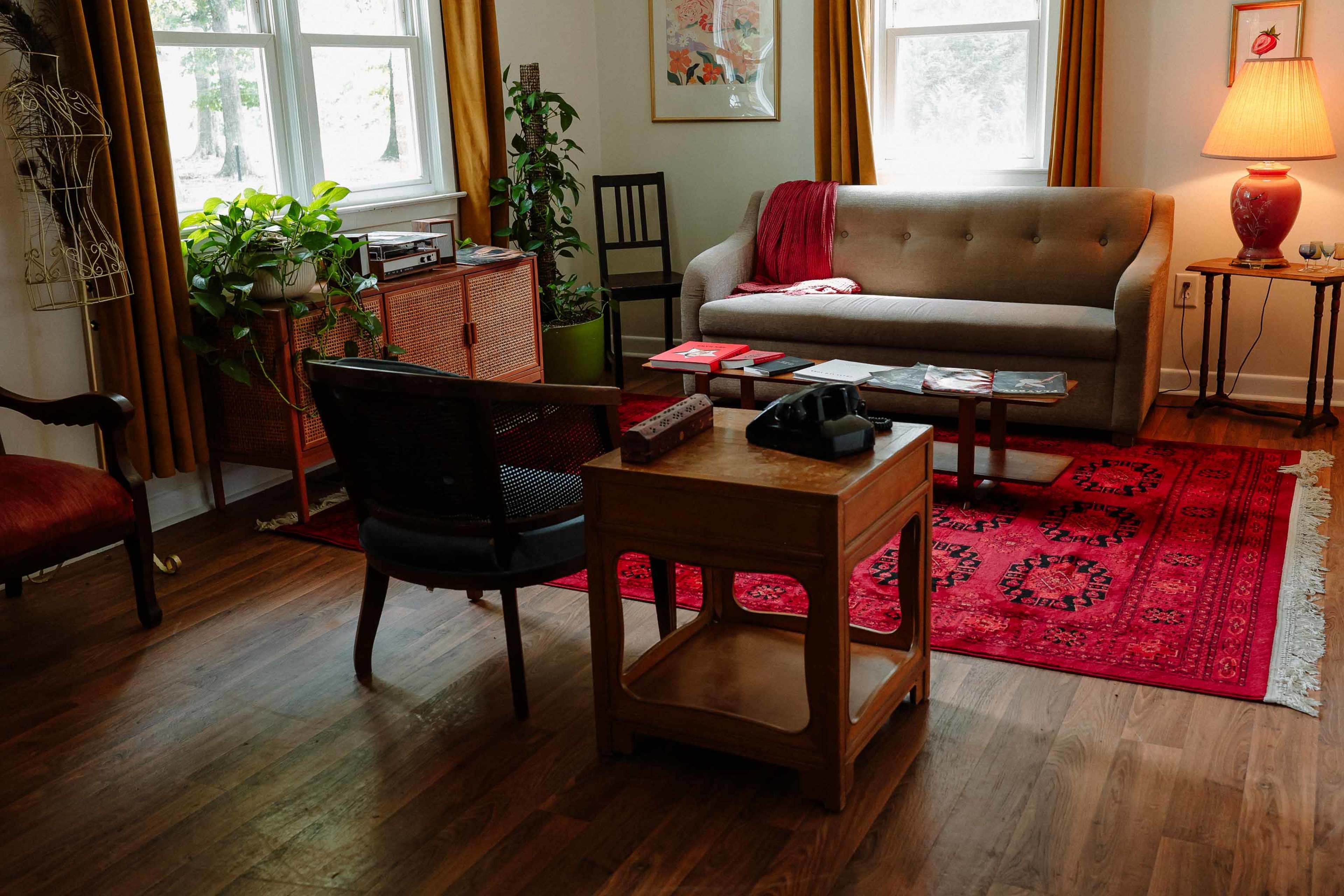 The image shows a cozy living room featuring a gray sofa, a wooden coffee table, a red rug, and plants near a large window.