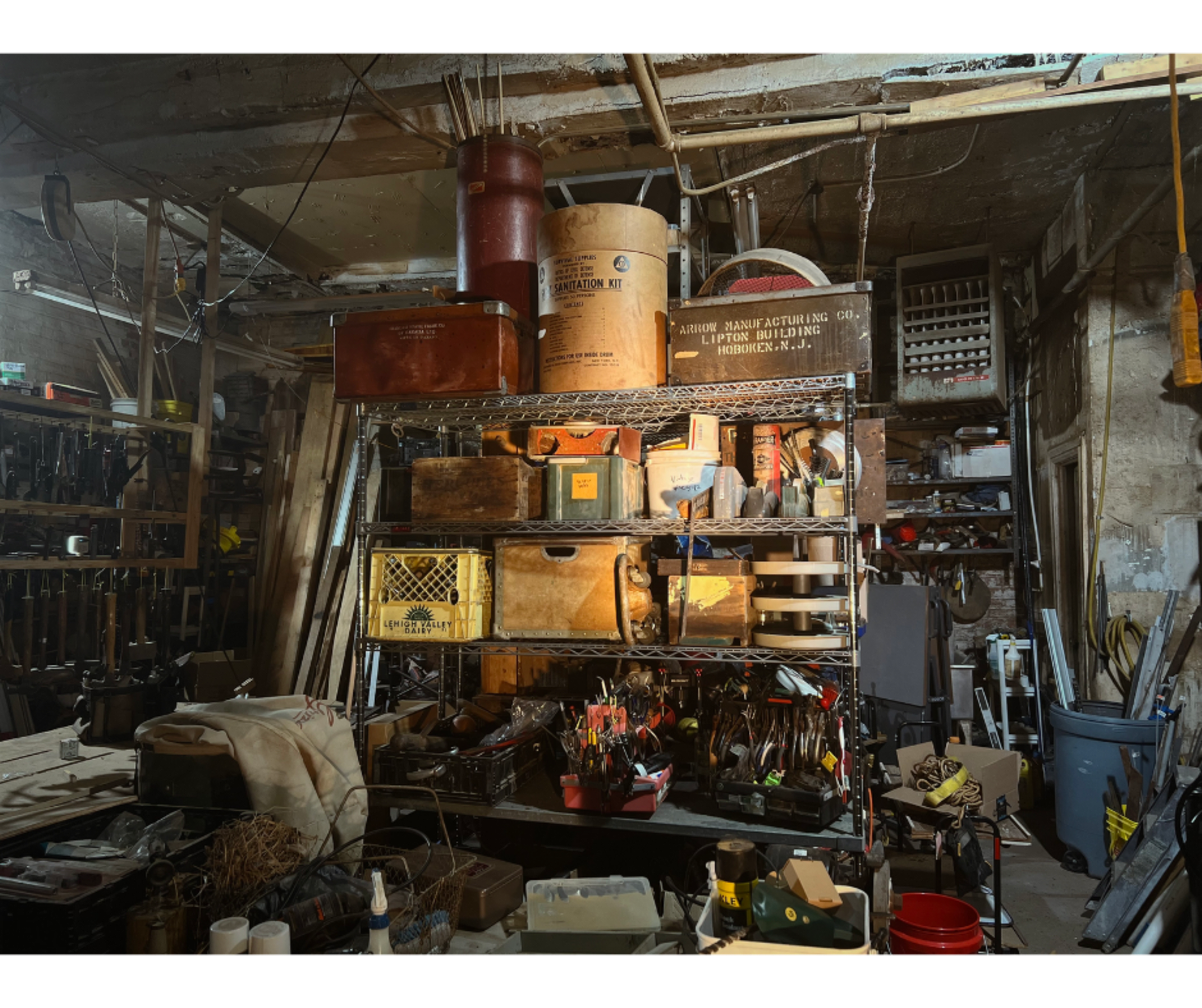The image shows a cluttered workshop filled with various tools, containers, and shelving units against a dimly lit background.