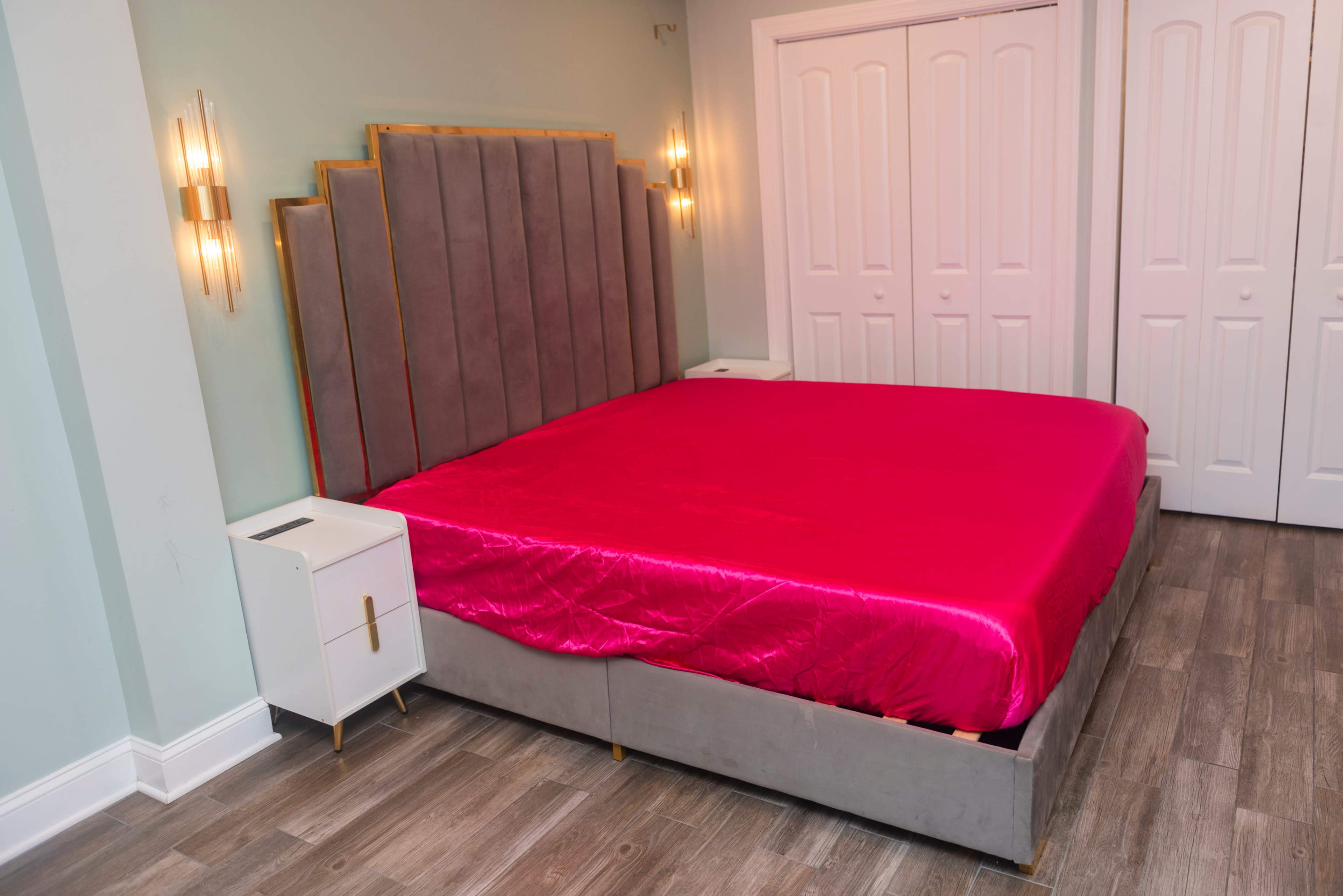 The image shows a modern bedroom with a large bed featuring a pink satin cover and a gray upholstered headboard, accompanied by a white nightstand beside it and double doors in the background.