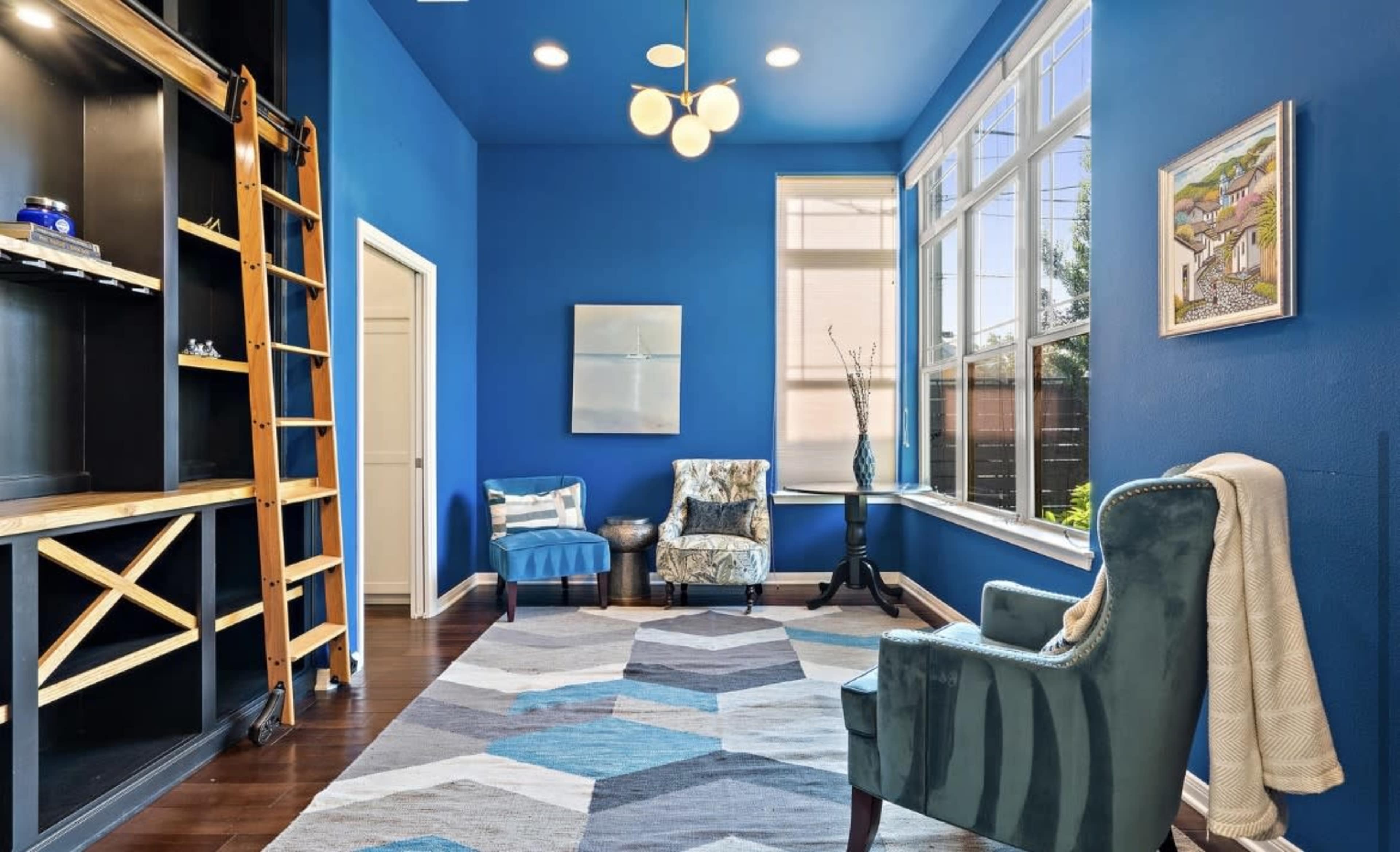 A bright room features blue walls, a geometric rug, two upholstered chairs, and a ladder against a shelving unit.