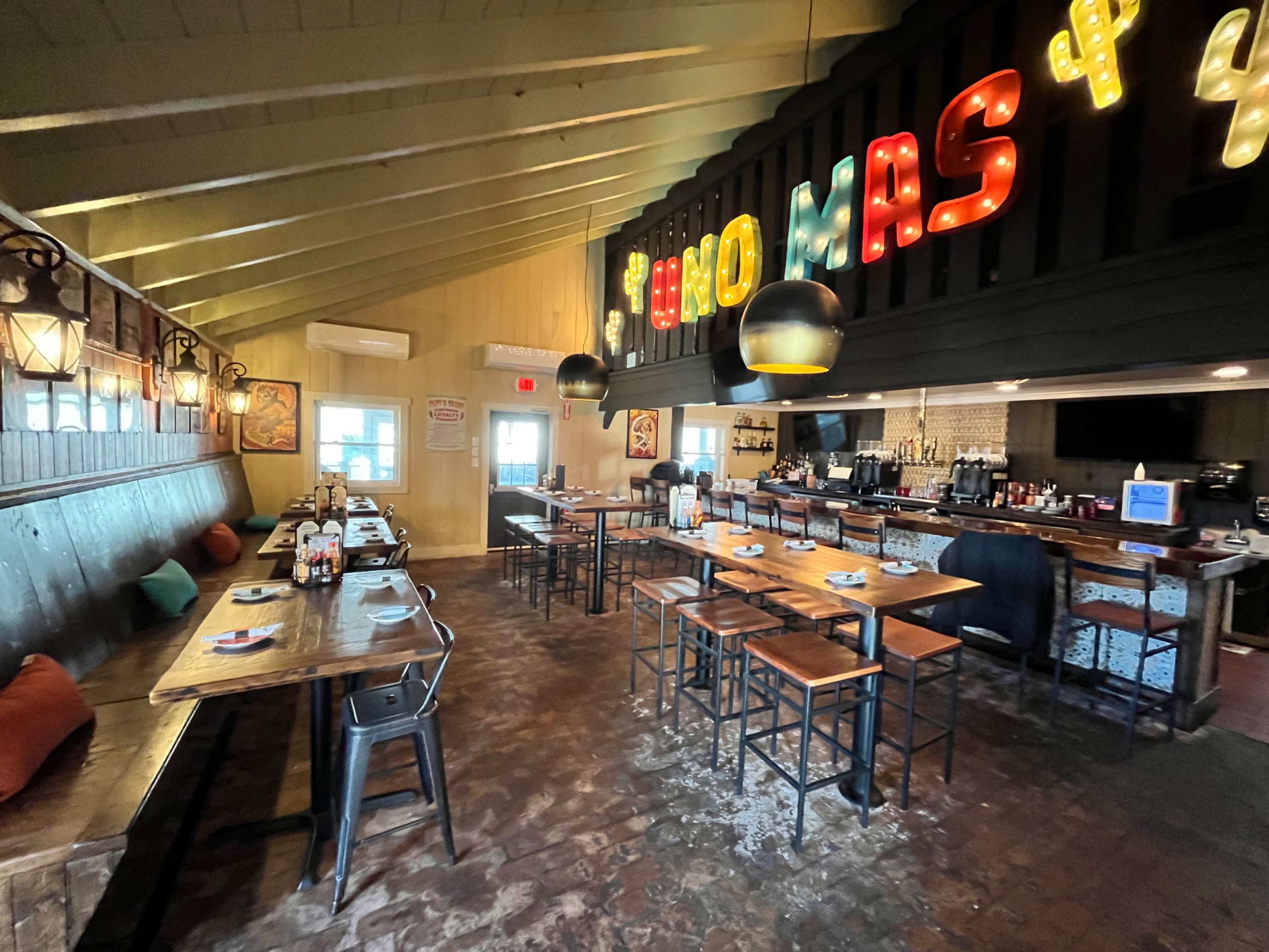 An interior of a restaurant with wooden tables, chairs, and a bar area illuminated by colorful lights.