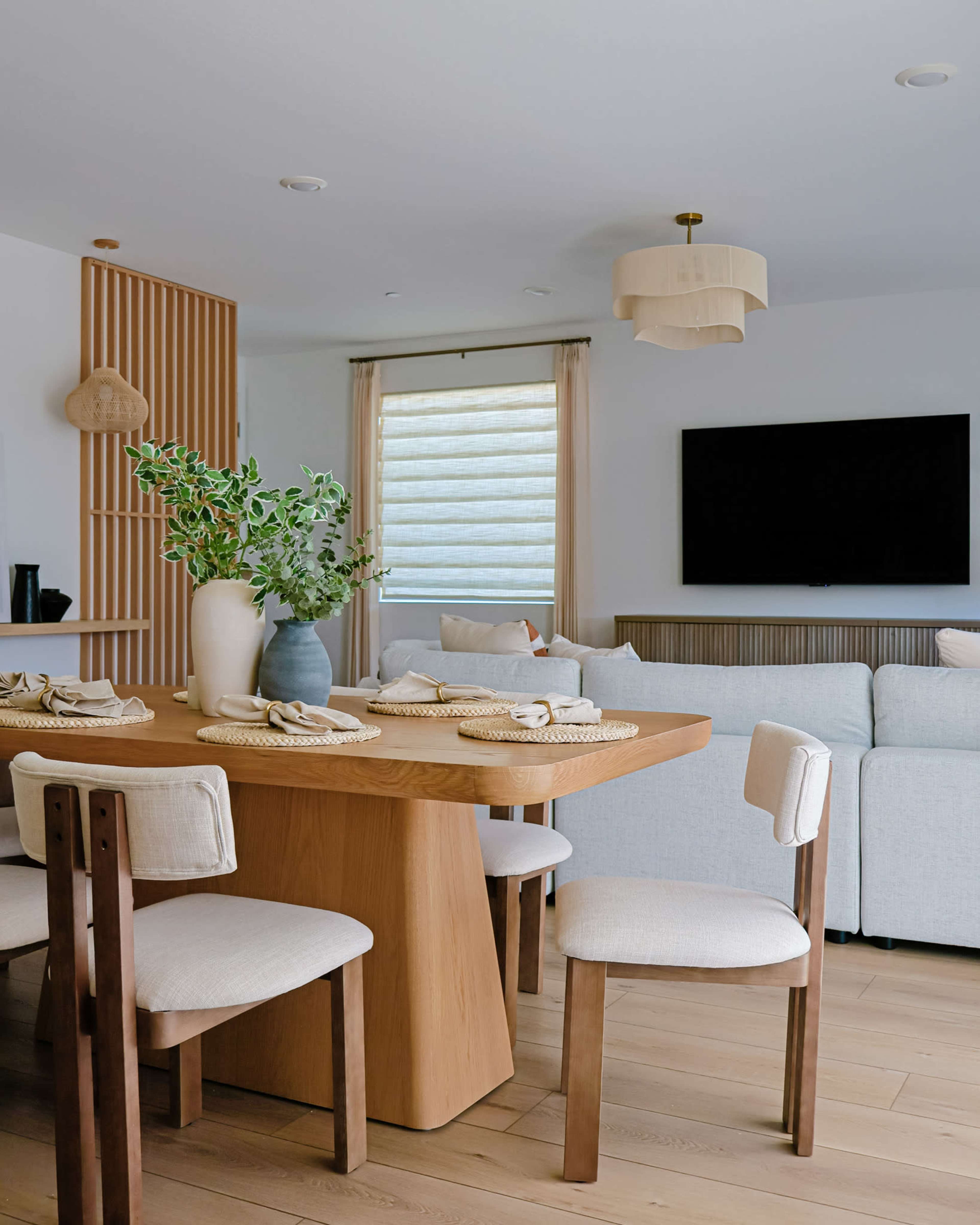 A modern dining area features a wooden table with white chairs, a vase with greenery, and a soft-colored sectional sofa next to a wall-mounted television.