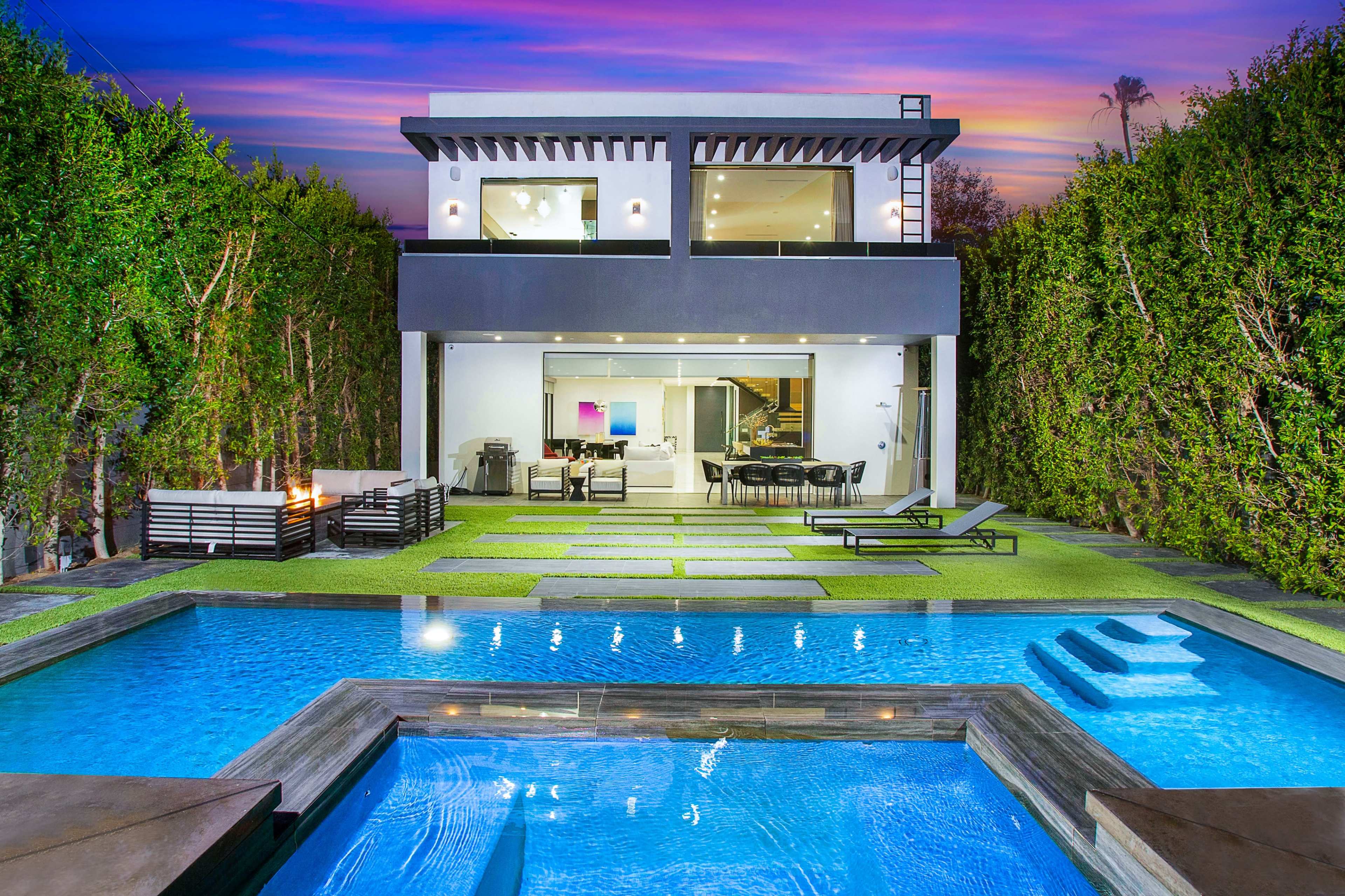 A modern two-story house with large windows overlooks a swimming pool surrounded by stone pathways and manicured grass.