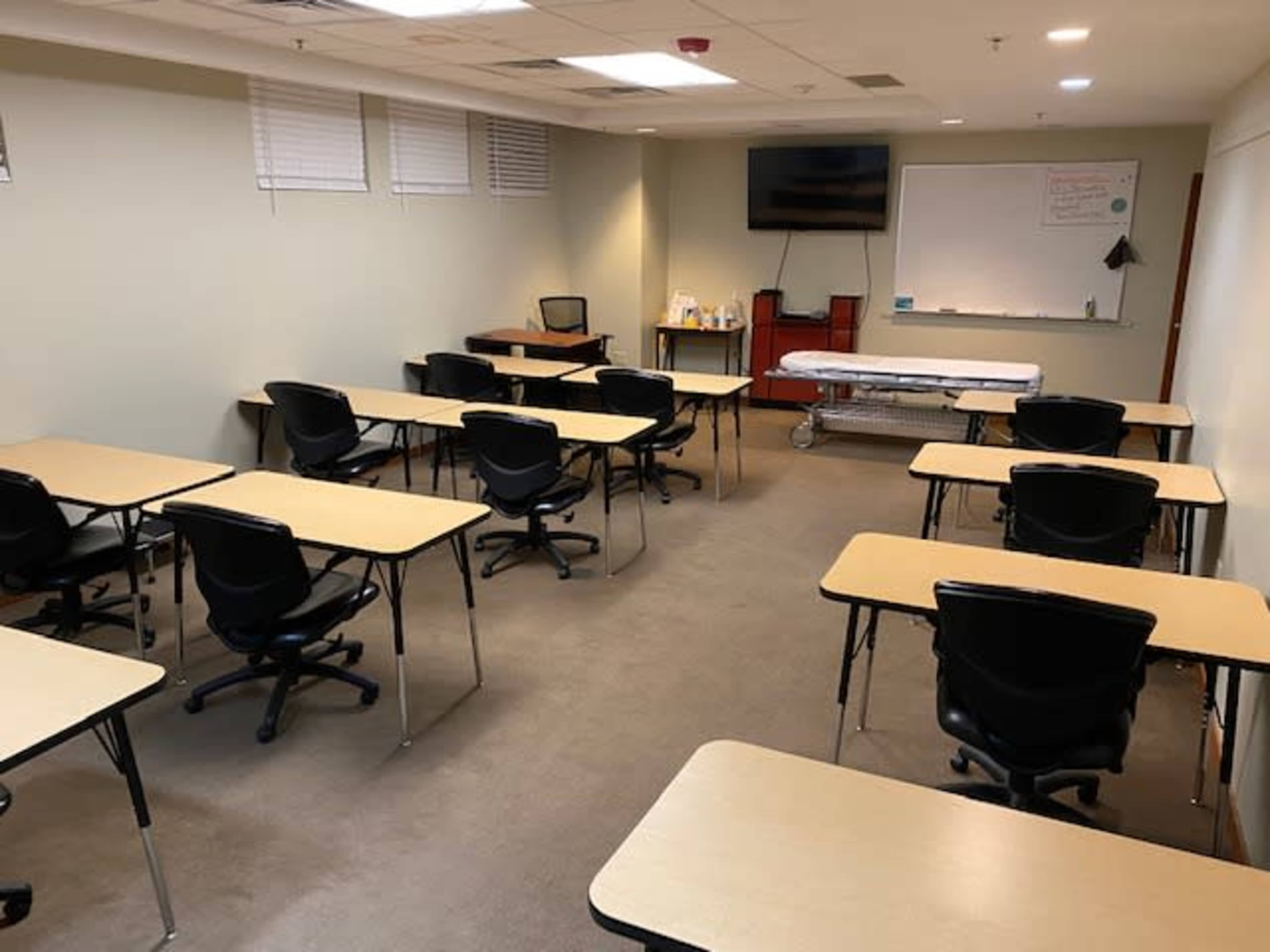 A sparsely furnished classroom with multiple empty tables and chairs arranged in rows, a whiteboard, and a television screen on the wall.