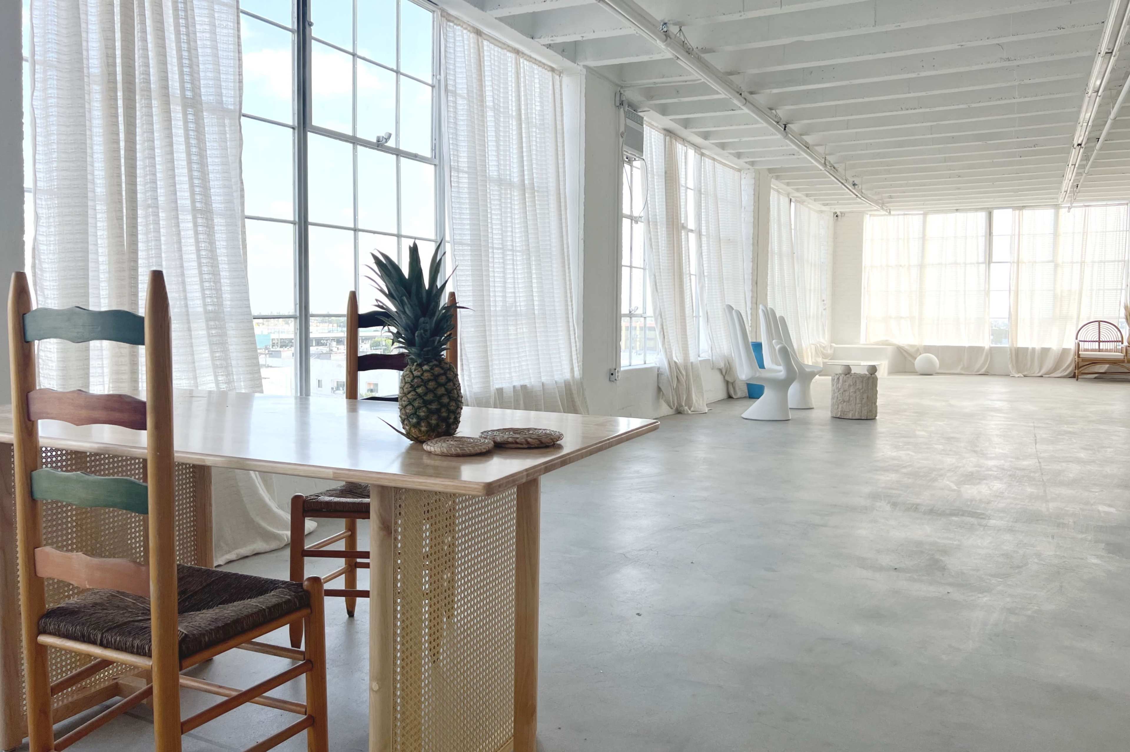 A bright, spacious room with large windows, minimal furniture including a table with chairs, and a pineapple as a centerpiece.