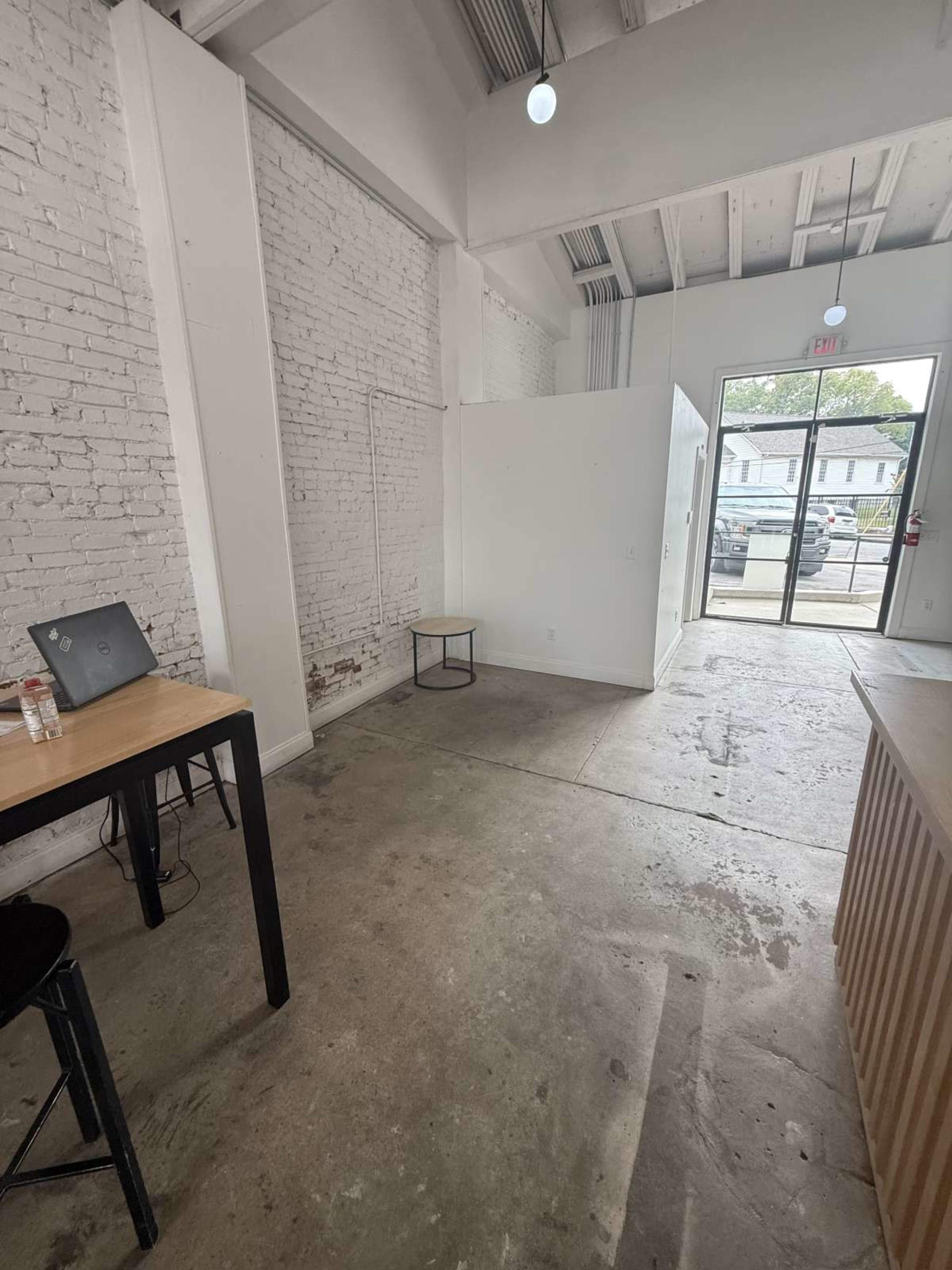 The image shows a sparsely furnished room with a concrete floor, a small table with a laptop, and a glass door leading outside.