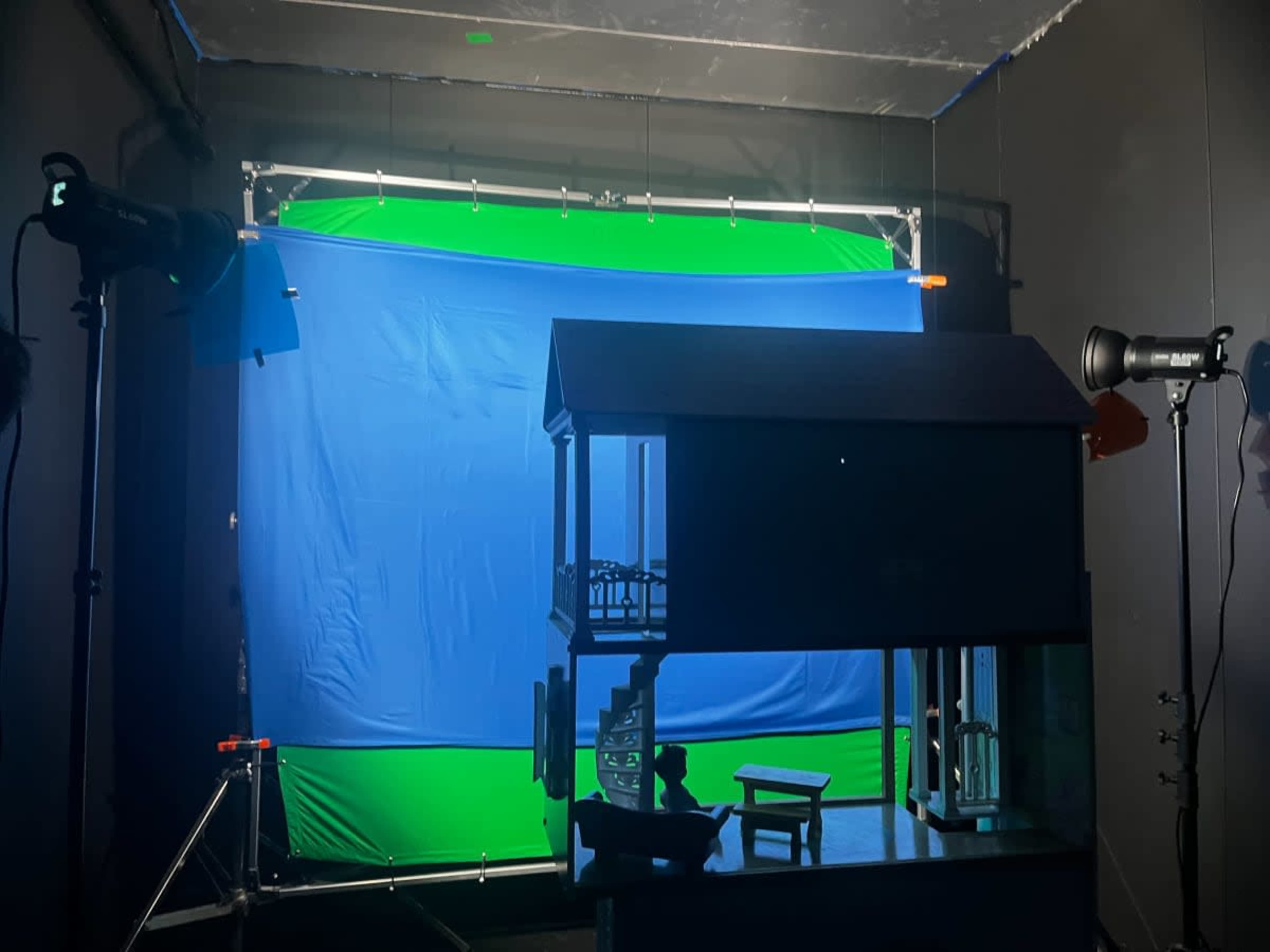 The image shows a small dollhouse positioned in front of a blue and green backdrop, with studio lights illuminating the scene.