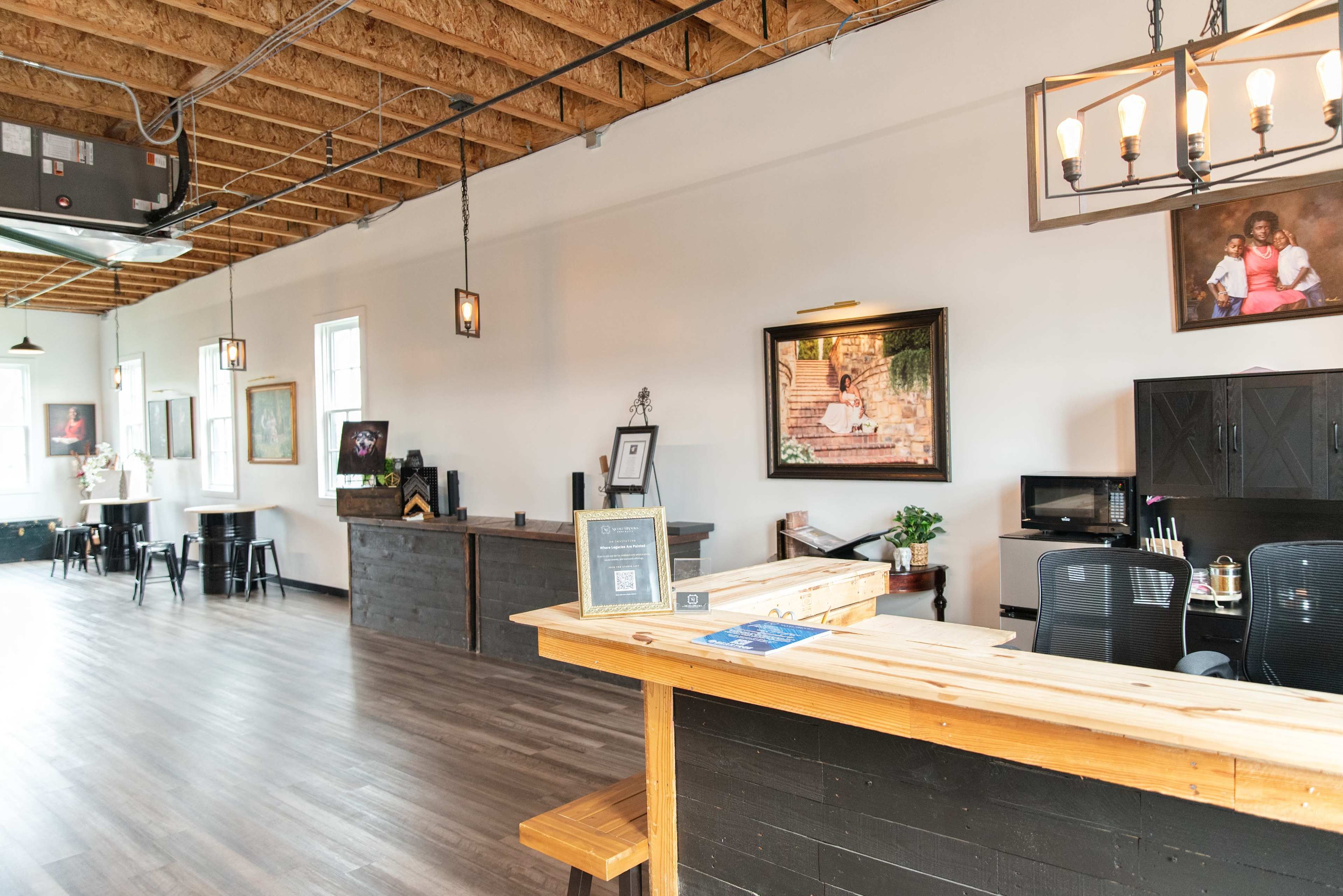 The image shows a modern, open interior space featuring a wooden reception counter, black office chairs, artwork on the walls, and a few tables in a well-lit setting.